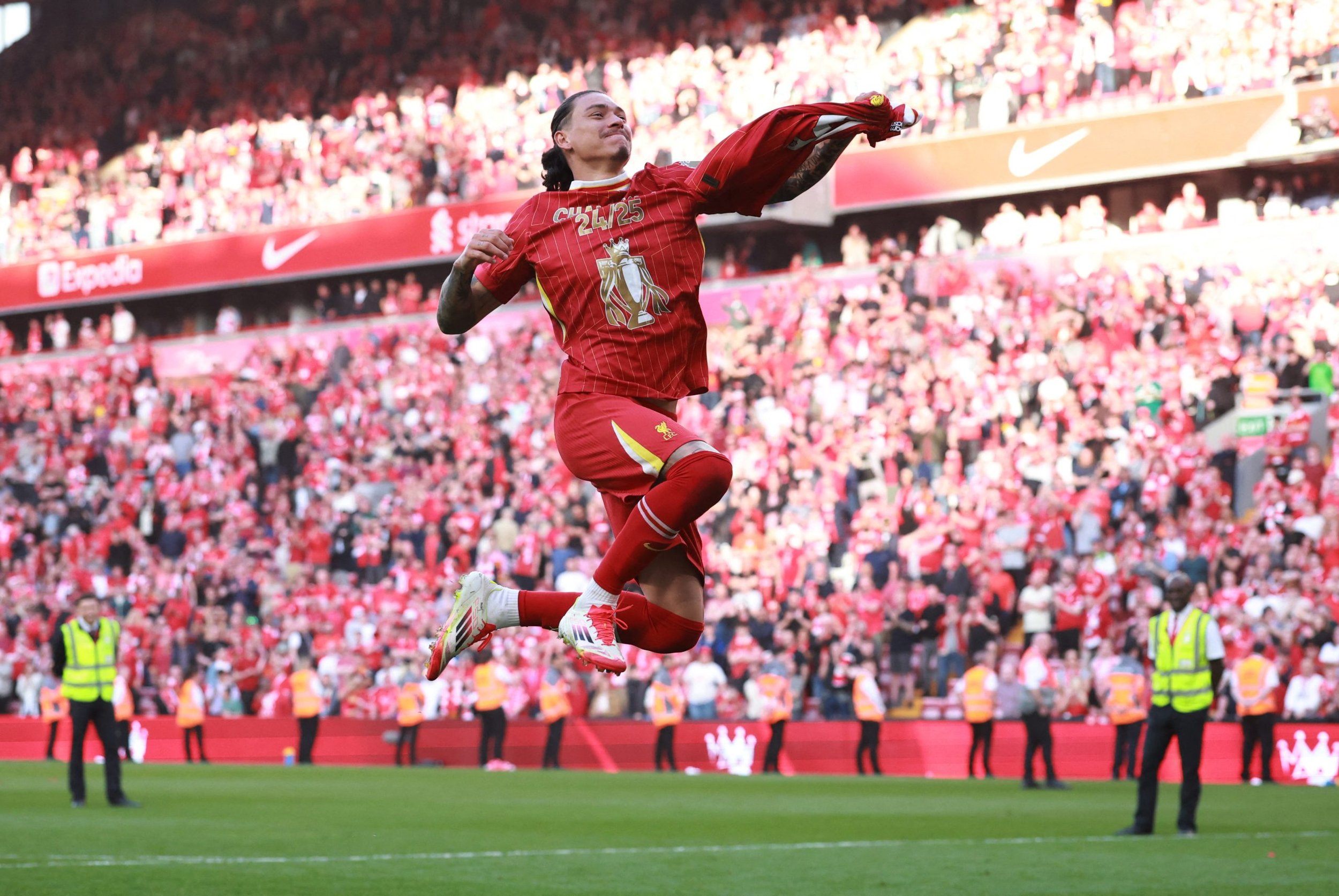 Soccer player jumping in celebration on a field, holding a red shirt with '25' on it, with a blurred crowd in the background wearing red. Security personnel in yellow vests are visible along the sidelines.