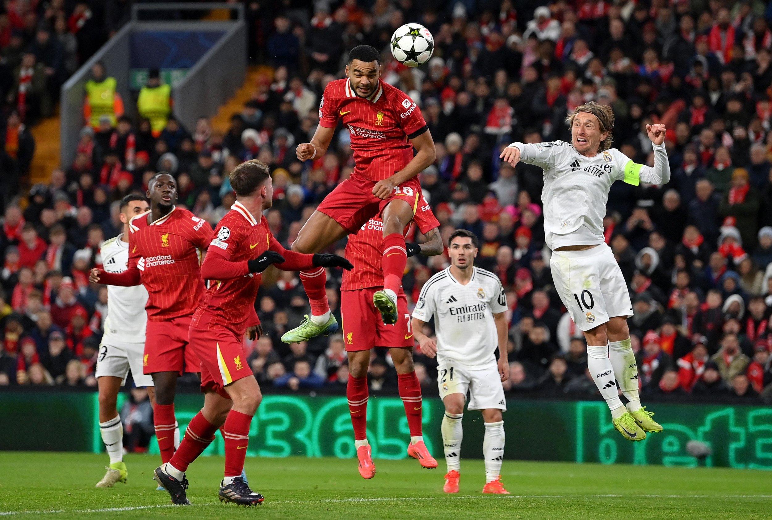Soccer players from Liverpool and Real Madrid jump for the ball during a match, with a crowd in the background.