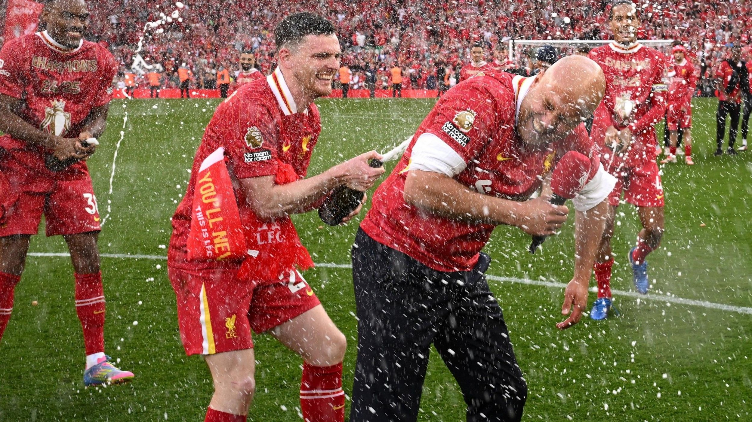 Football players celebrating with champagne on a field, wearing red uniforms; a crowd in the background.