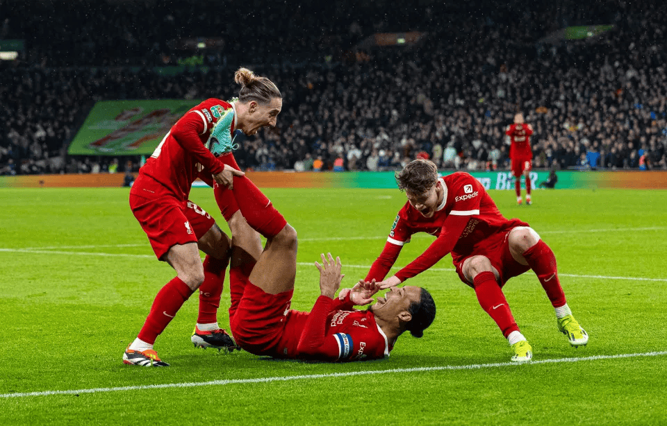 Soccer players from a team in red uniforms celebrating on the field, with one player lying on the ground smiling and others leaning over him. A large crowd is visible in the background.