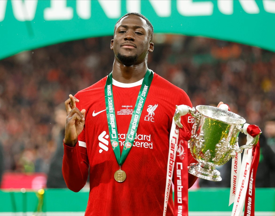 Soccer player wearing a red Liverpool FC jersey, holding a trophy with white ribbons labeled 'Carabao Cup,' and a green winner's medal around his neck.