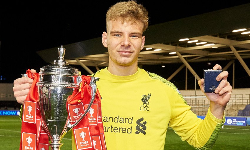 A soccer player in a yellow jersey holding the FA Youth Cup trophy and a medal. The trophy is silver with red ribbons labeled "The FA." The player is standing in a stadium with seating and a roof structure visible.
