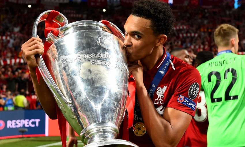 Soccer player kissing the UEFA Champions League trophy in a stadium, wearing a red jersey, surrounded by teammates and medallions.