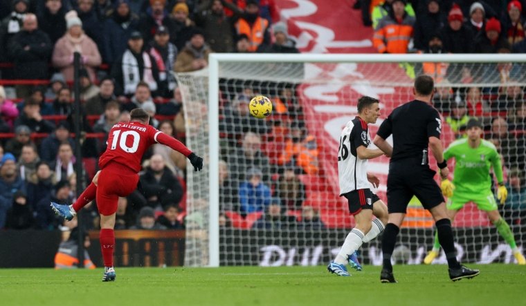 A soccer player in a red jersey, number 10, kicking a ball during a match. Opposing team players and a referee are visible in the background. Spectators watch from the stands.