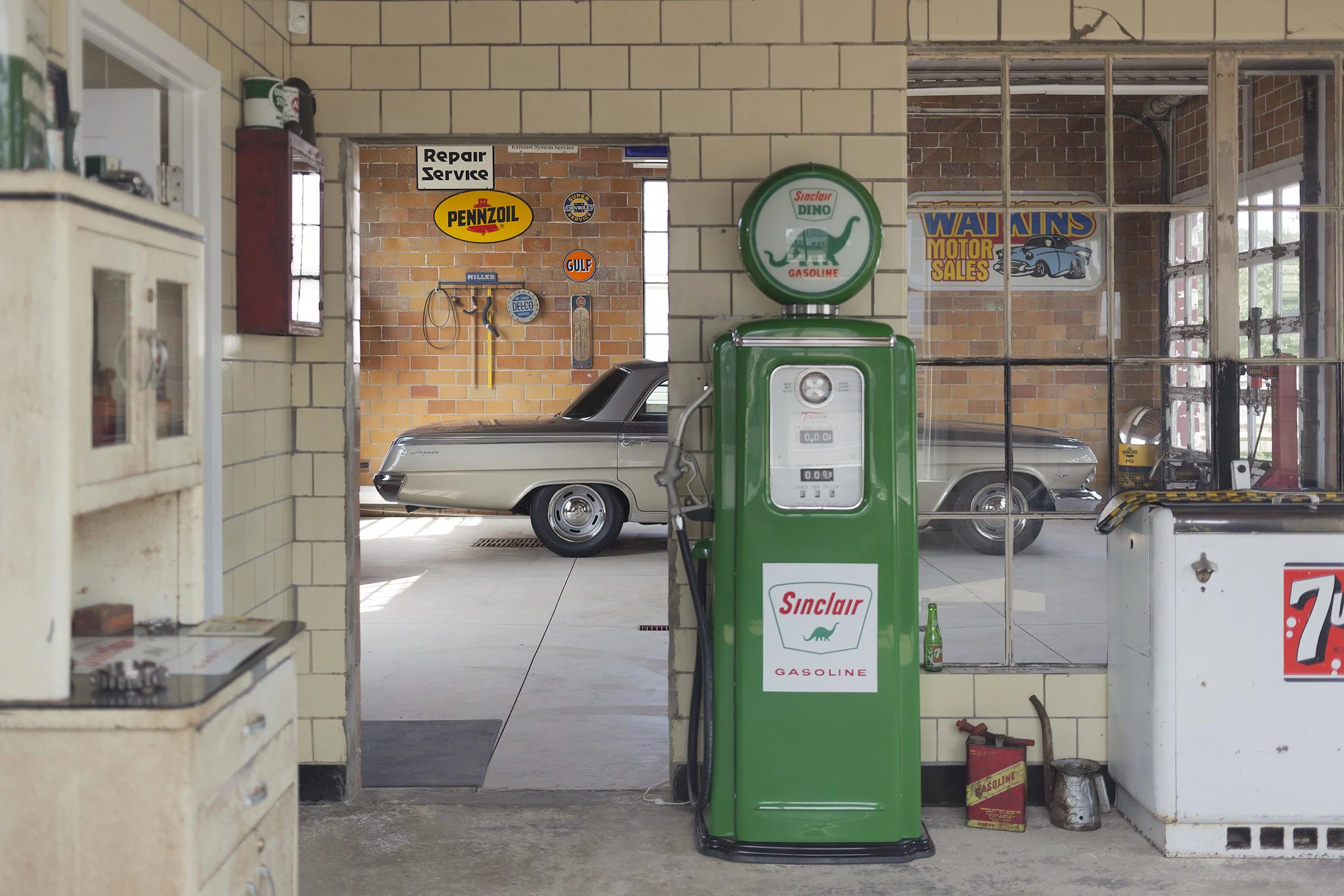 Interior of a vintage gas station, featuring a green Sinclair gasoline pump in the foreground, classic cars parked inside, and retro signs on the brick walls, including a Walkins Motor Sales banner and various oil company logos.