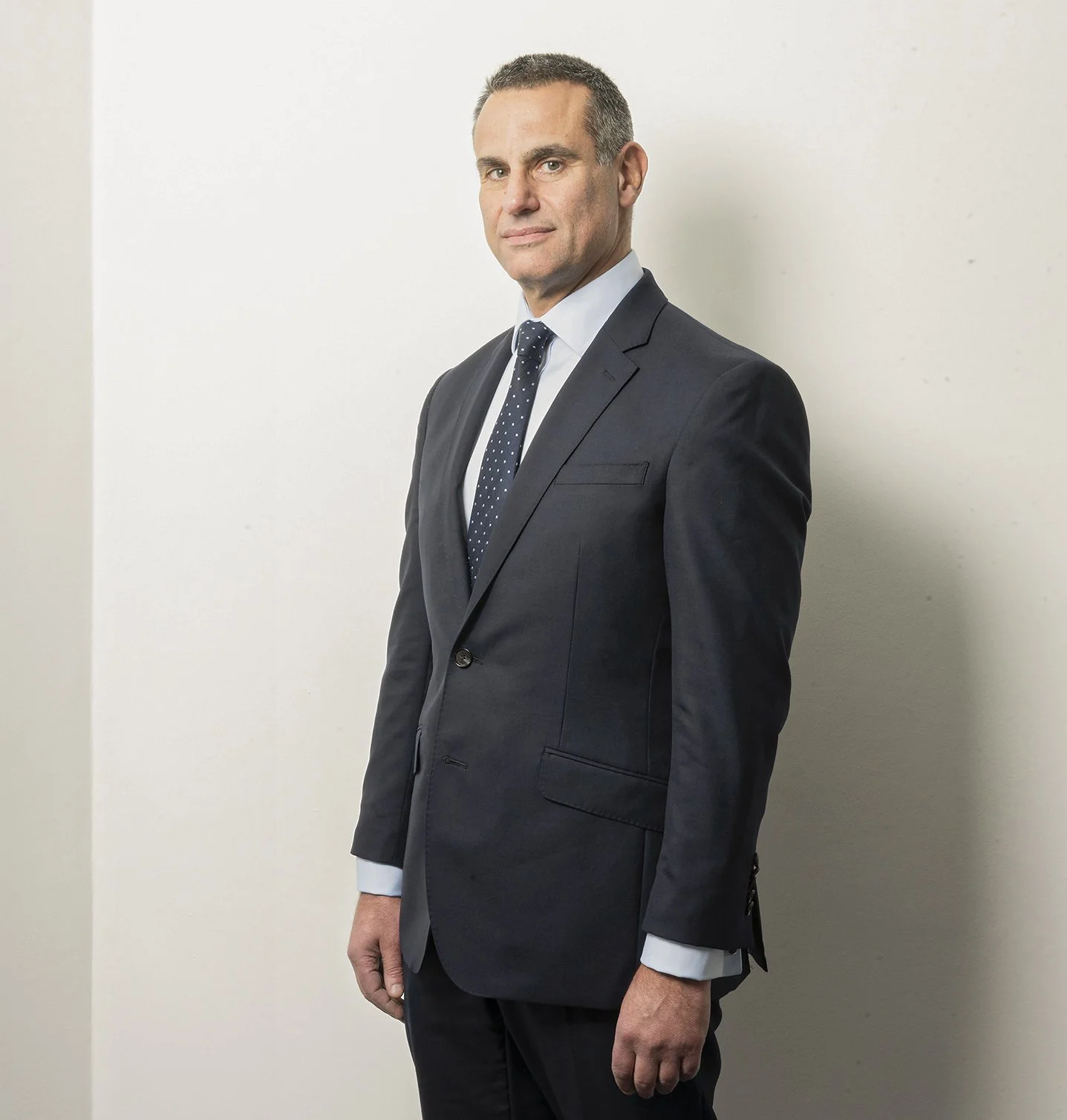 A man in a dark suit, white shirt, and dotted tie, standing against a plain white wall with a confident expression.