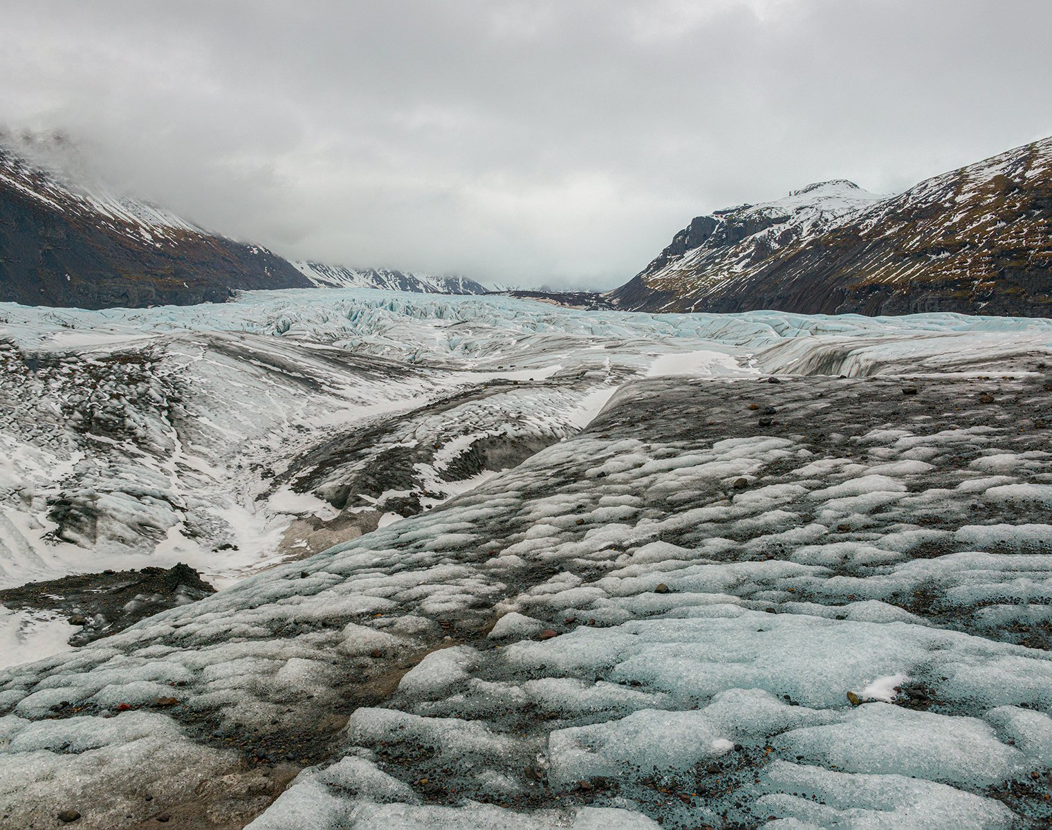 A glacier with ice and snow in a valley surrounded by mountains, under a cloudy sky.