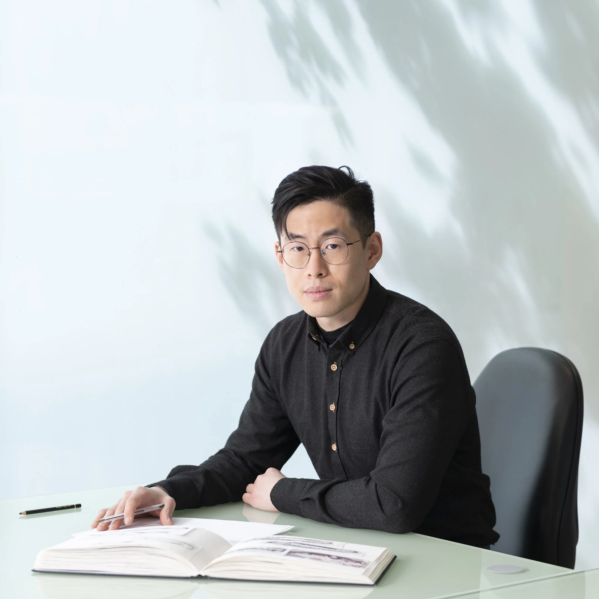 A young man wearing glasses, a black button-up shirt, sitting at a white desk with an open scrapbook and a pen, in front of a light-colored wall with shadows of leaves.