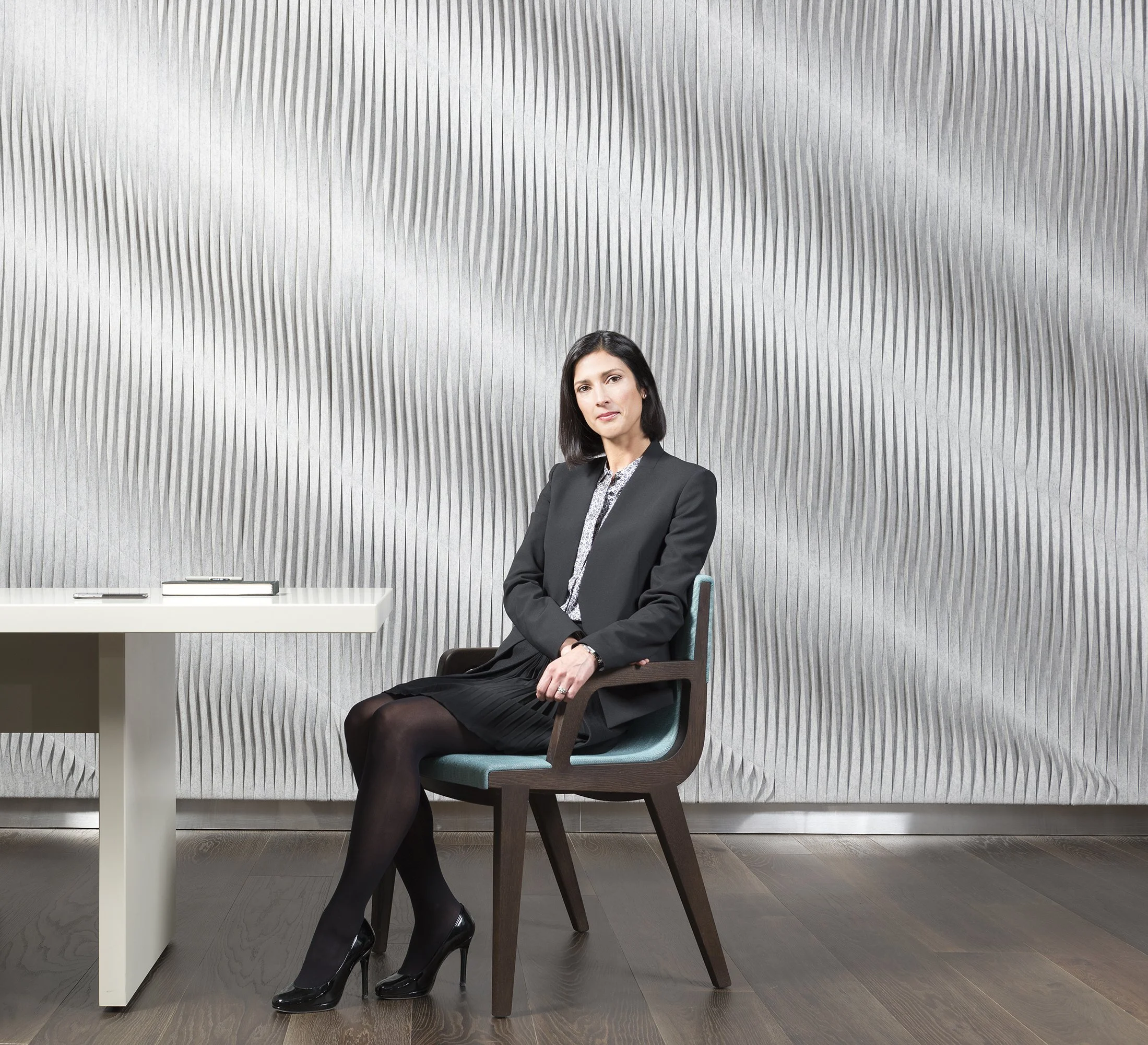 A woman with dark hair wearing a black blazer and skirt, sitting on a chair in a modern office setting with a textured gray wall in the background and a white table with a notebook and phone.