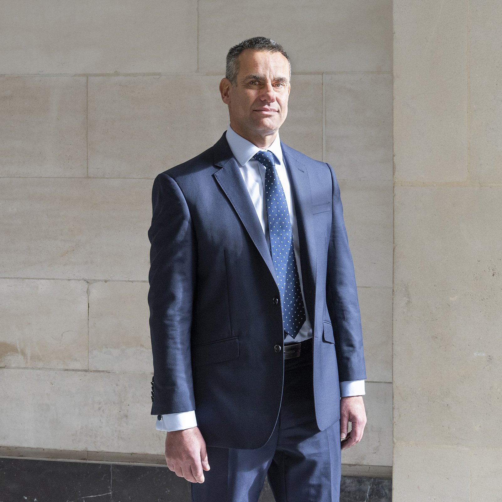 A confident middle-aged man dressed in a navy blue business suit, white shirt, and navy polka dot tie standing against a beige stone wall.