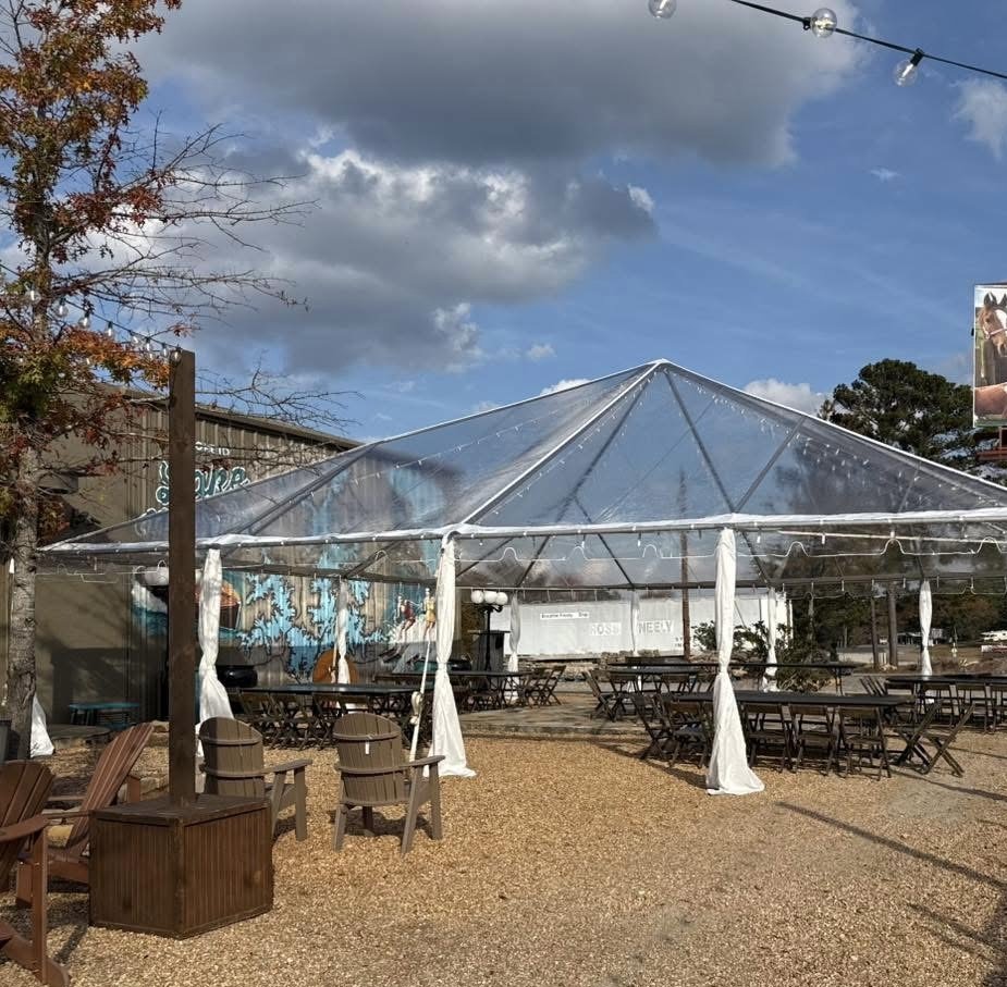 Empty outdoor seating area with wooden chairs and tables under a large clear tent, with string lights overhead, trees with fall foliage, and a partly cloudy sky.
