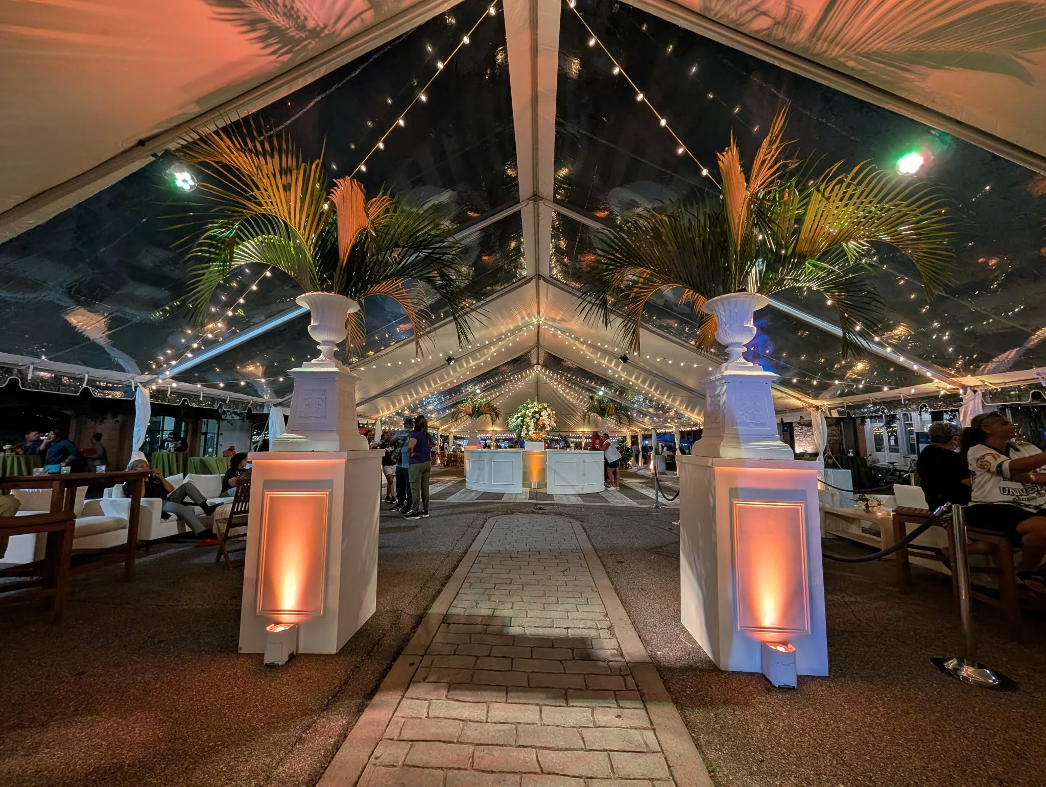 Elegant event tent with string lights, large floral arrangements in white vases, palm leaves, and guests seated at tables and couches, illuminated by warm uplighting.