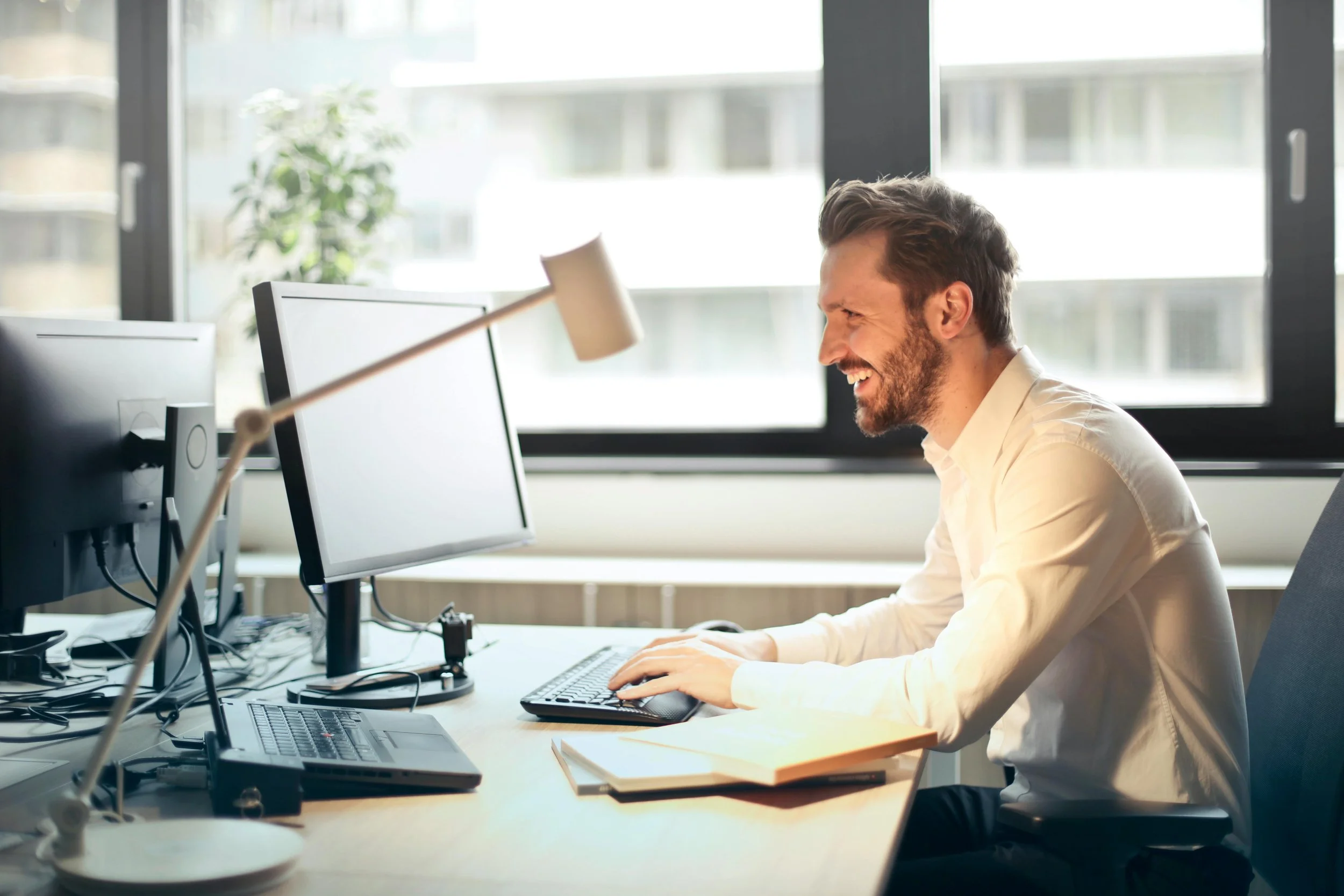 Hombre trabajando en una oficina con computadora y lámpara de escritorio, sonriendo mientras usa el teclado.