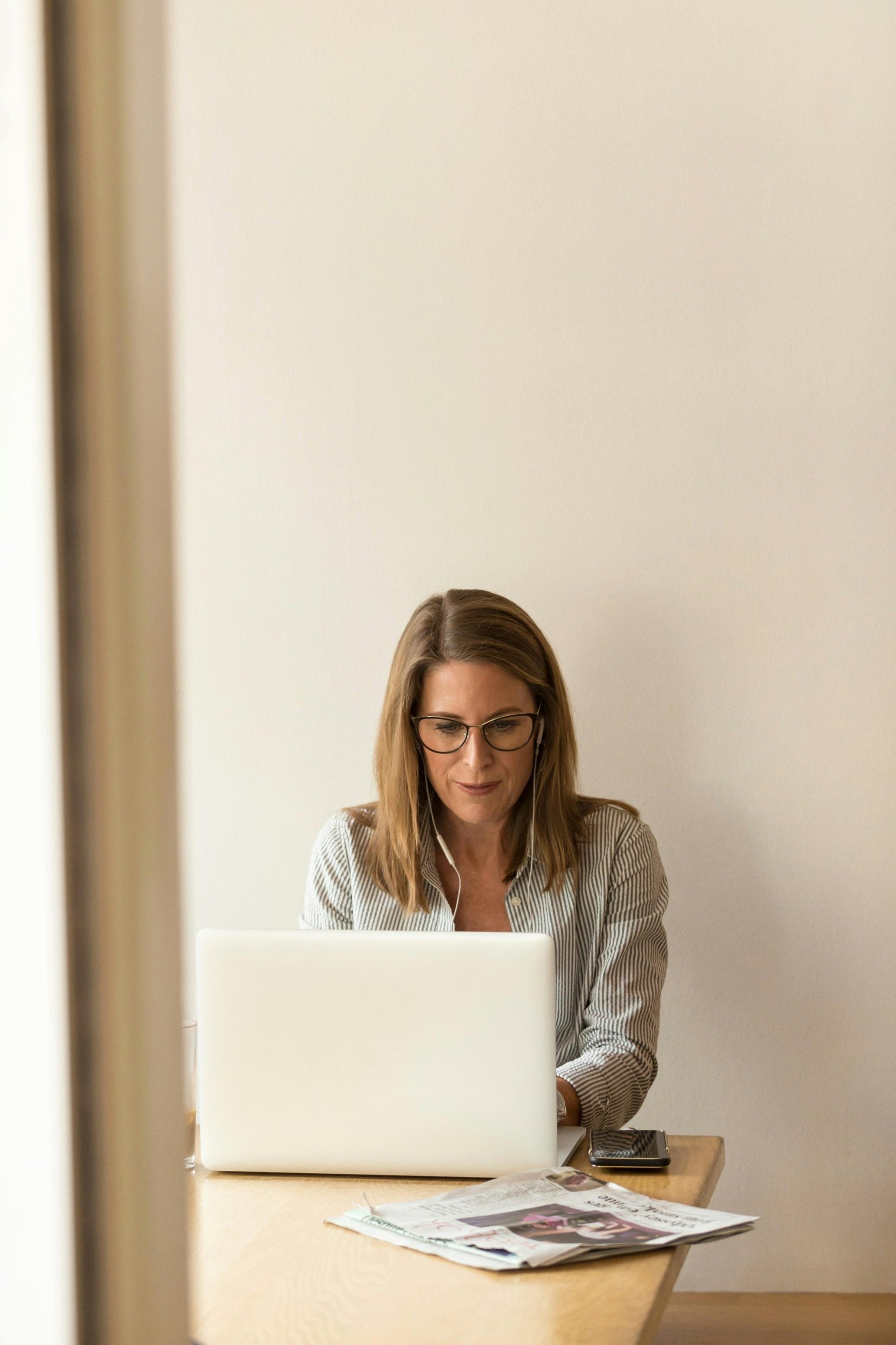 Persona trabajando en una computadora portátil sobre una mesa con periódicos, usando gafas y auriculares, en un ambiente interior.