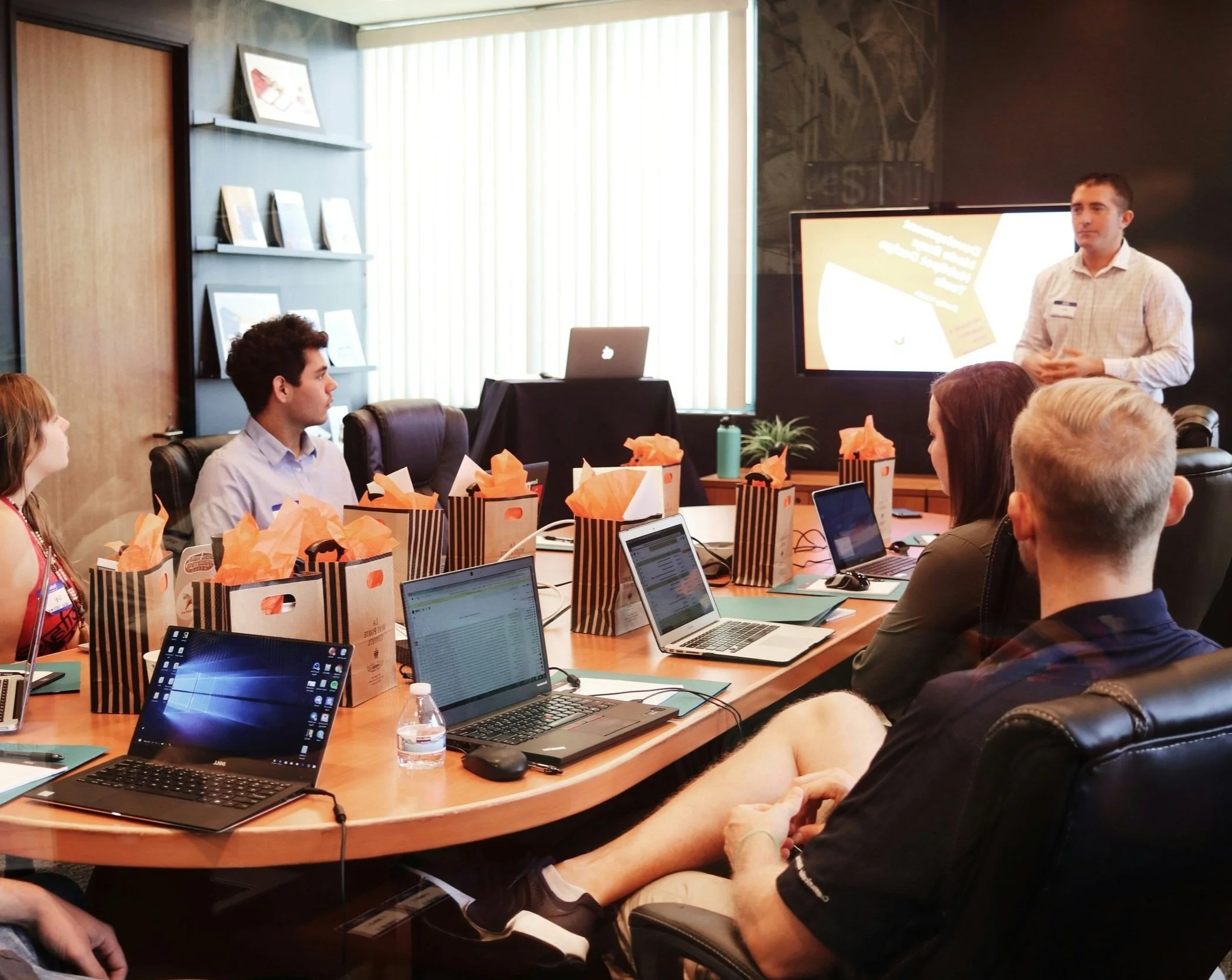 Personas en una reunión de negocios en una sala de conferencias, con laptops y una presentación en una pantalla grande.