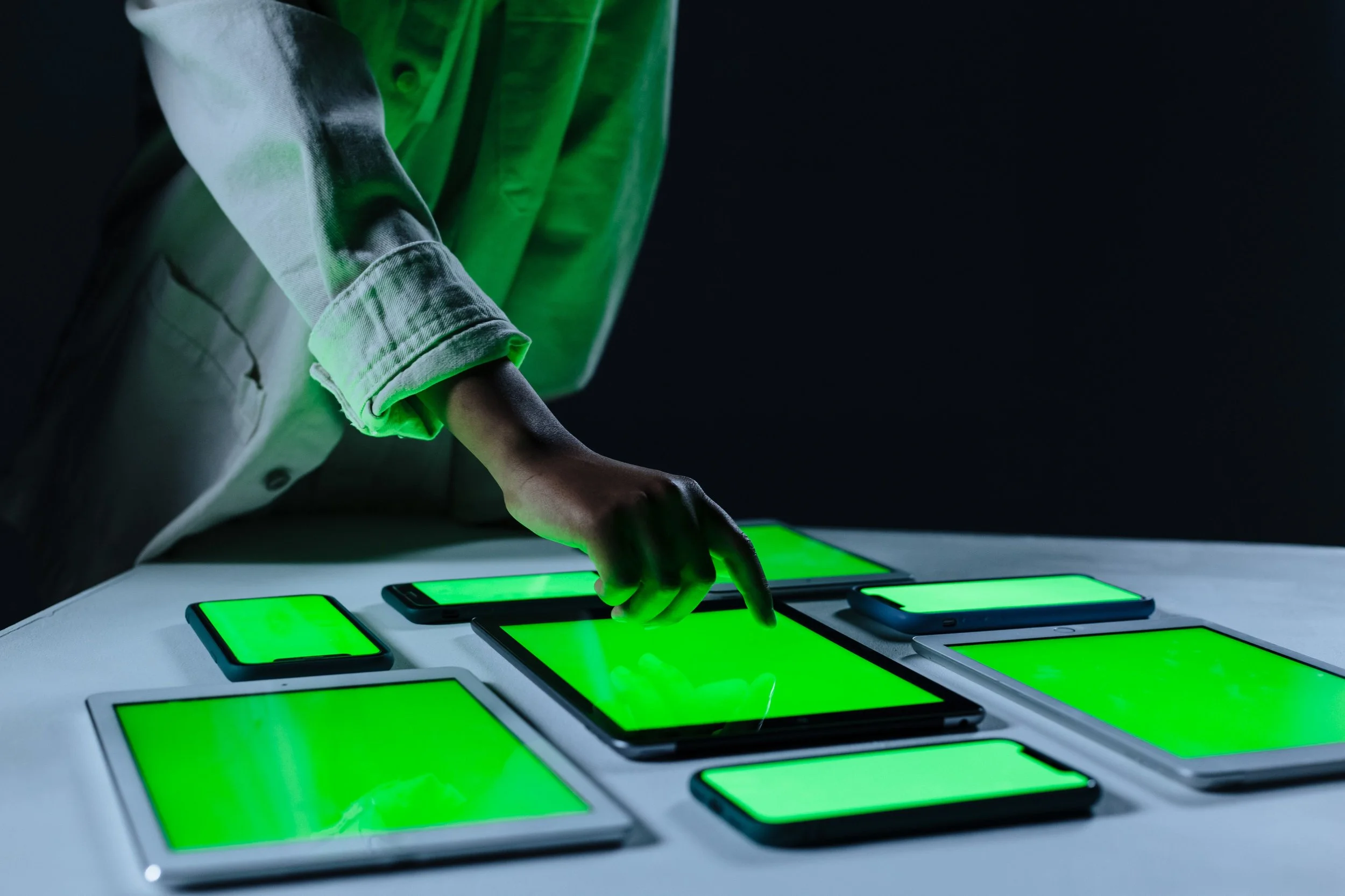 Manos de una persona tocando dispositivos electrónicos con pantallas verdes en una mesa, en un entorno oscuro.