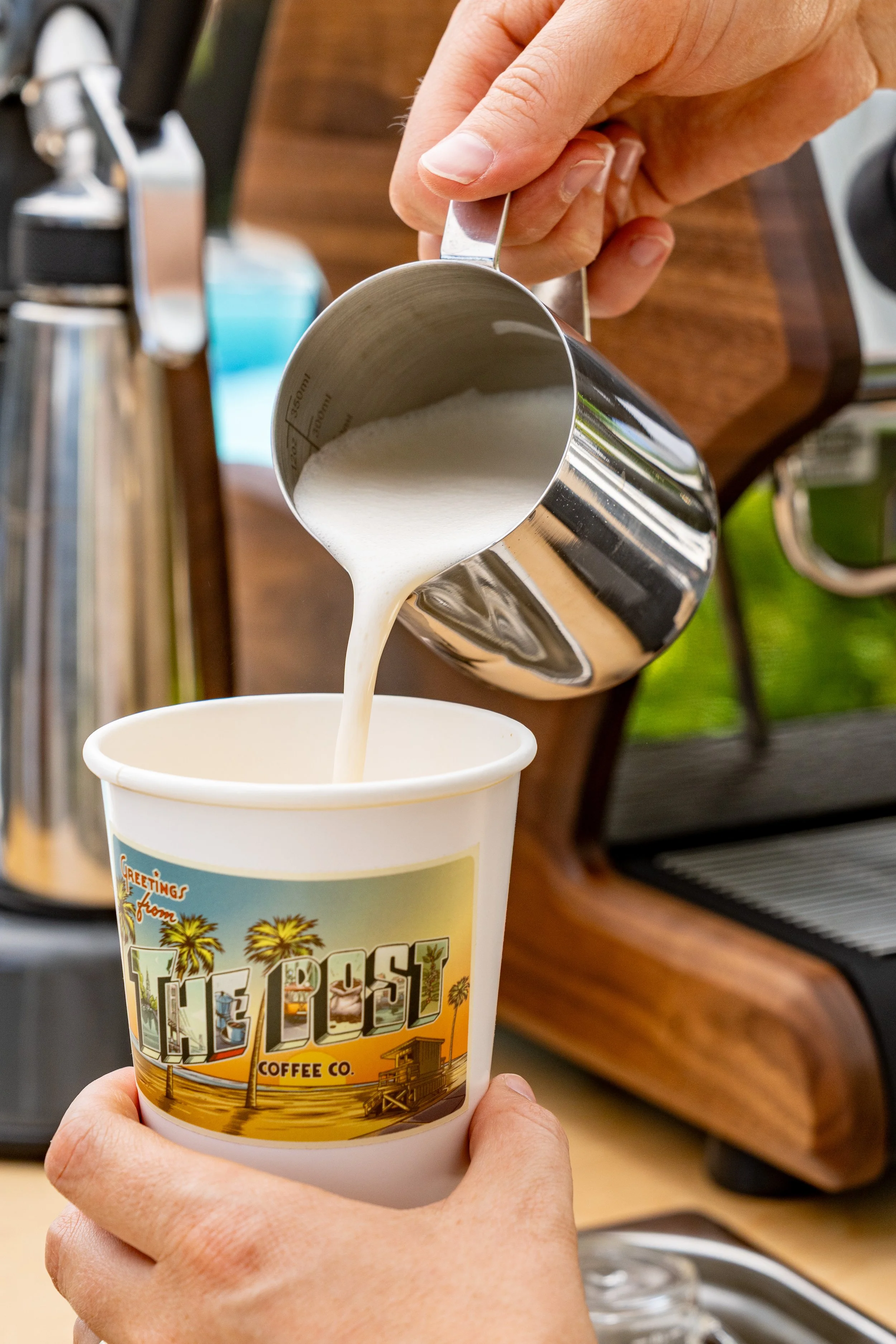 A person pours frothed milk from a metal pitcher into a paper coffee cup with a beach-themed design. The background includes a coffee machine.