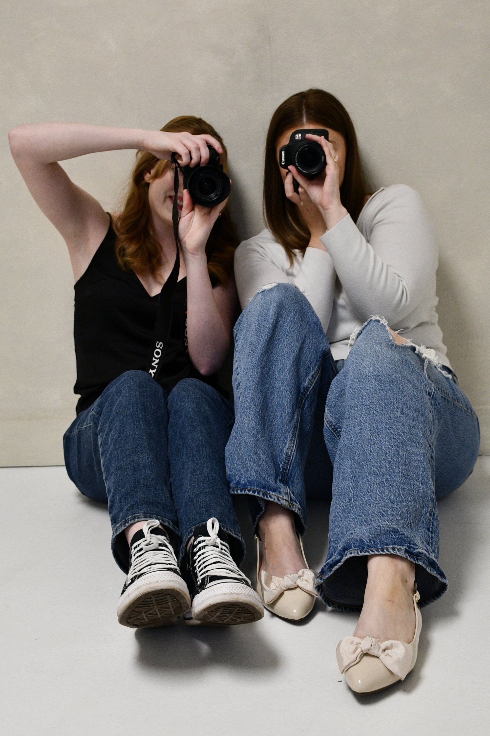 Two women sitting on the floor against a plain beige wall, holding cameras and taking a photo of themselves, wearing casual clothes and sneakers or ballet flats.