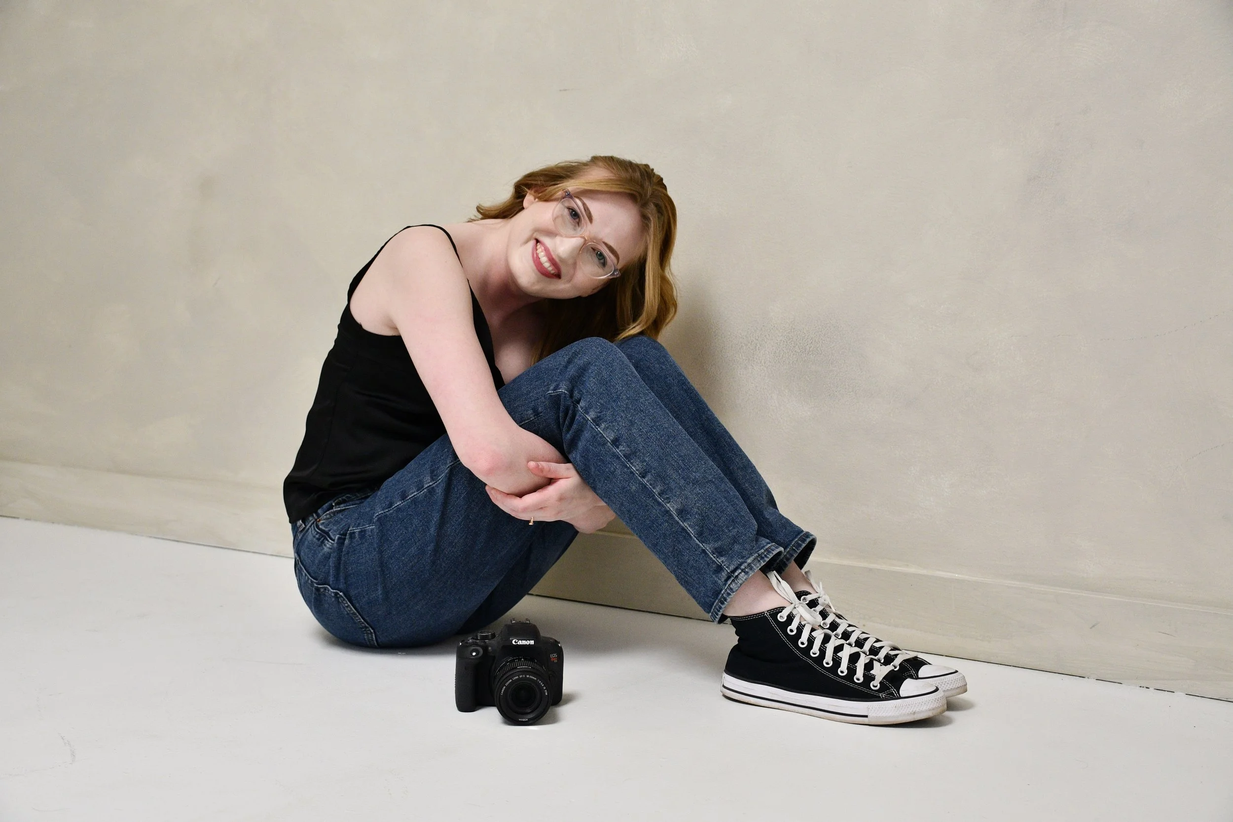 A smiling woman with red hair, glasses, and fair skin sits on the floor with her knees drawn up, wearing a black tank top, blue jeans, and black high-top Converse sneakers. A Canon camera is placed on the floor nearby, against a plain beige wall.