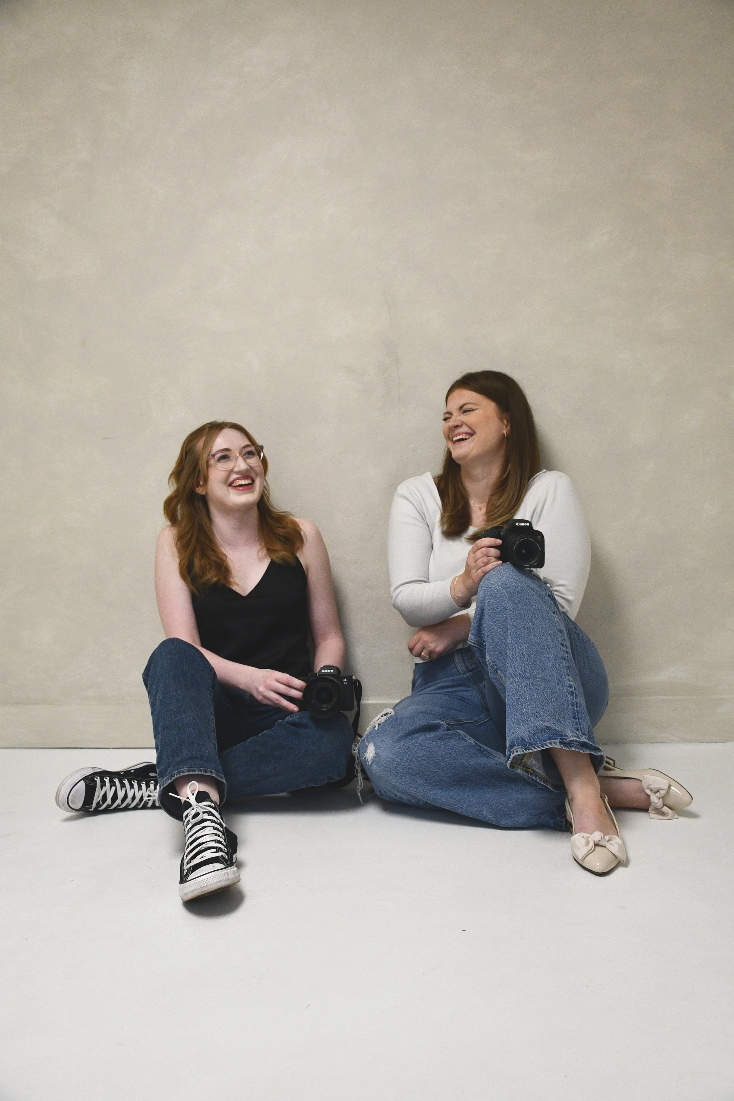 Two women sitting on the floor against a light-colored wall, smiling and laughing, each holding a camera.