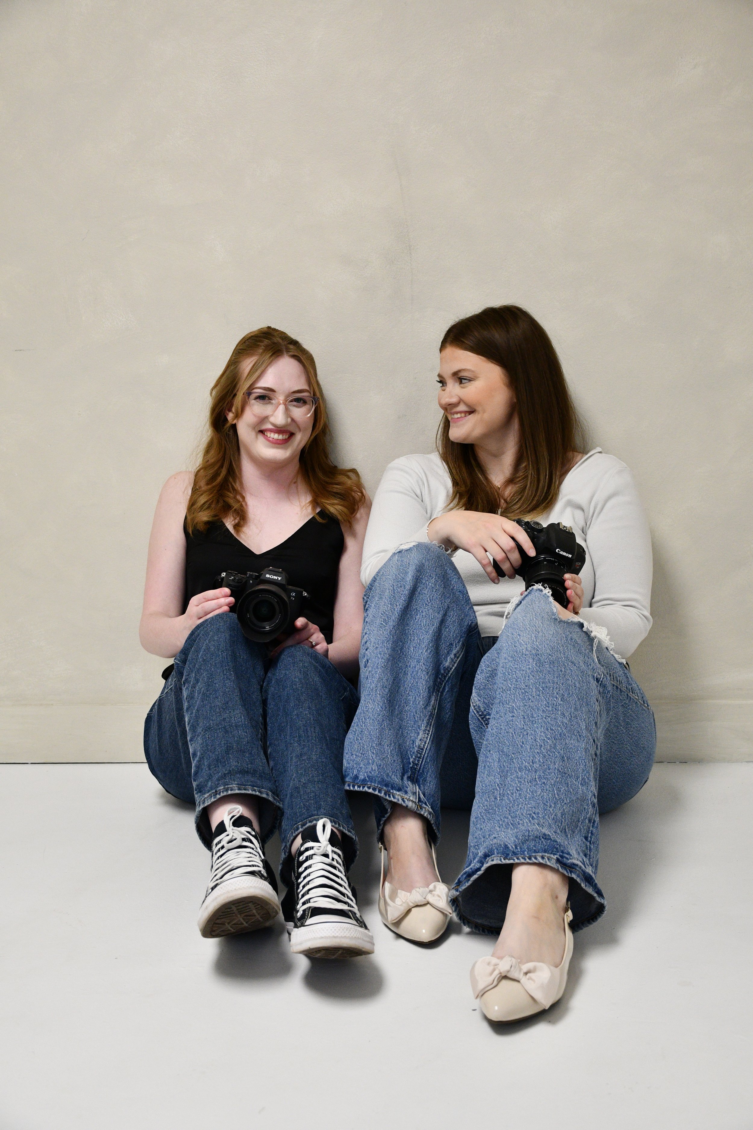 Two women sitting on the floor against a wall, holding cameras, smiling and looking at each other.