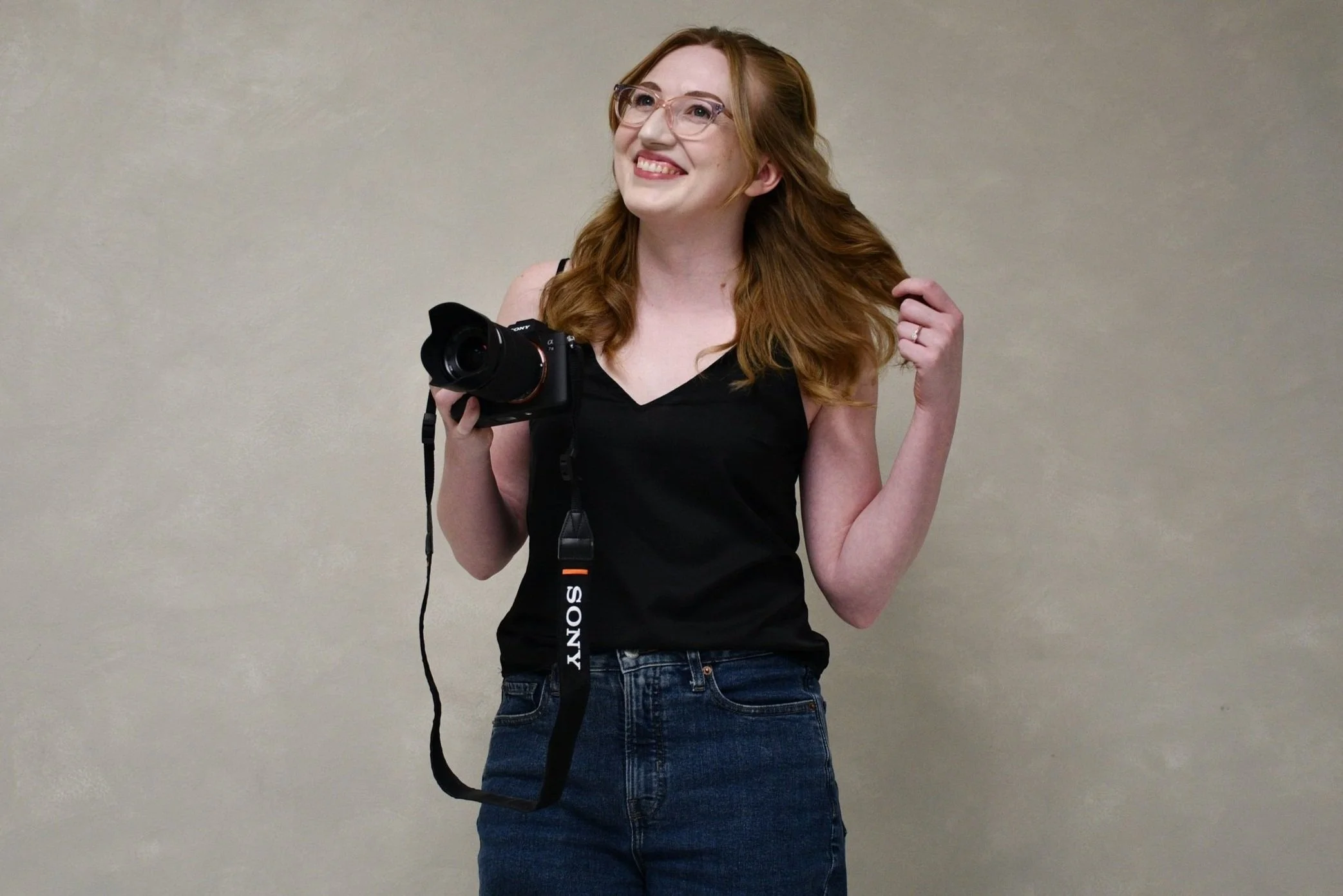 Young woman with red hair, glasses, black sleeveless top, and blue jeans holding a DSLR camera, smiling and touching her hair, standing against a plain beige wall.