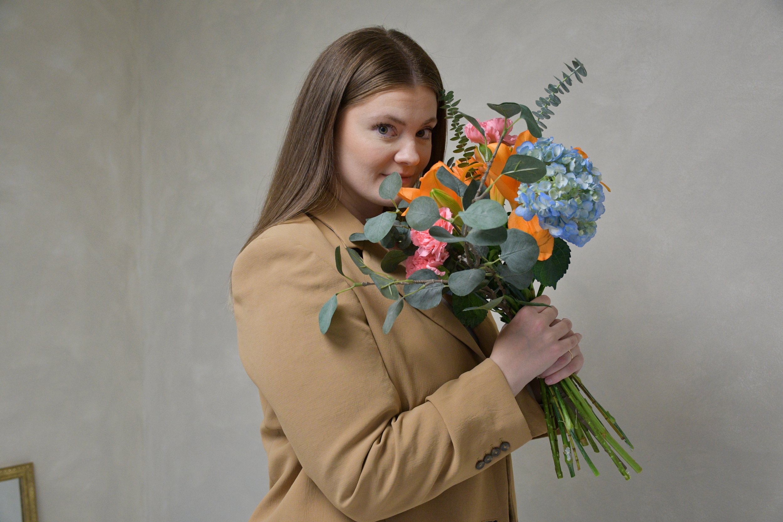 Woman with long brown hair holding a colorful bouquet of flowers, including pink, blue, and orange blooms, against a plain gray wall.