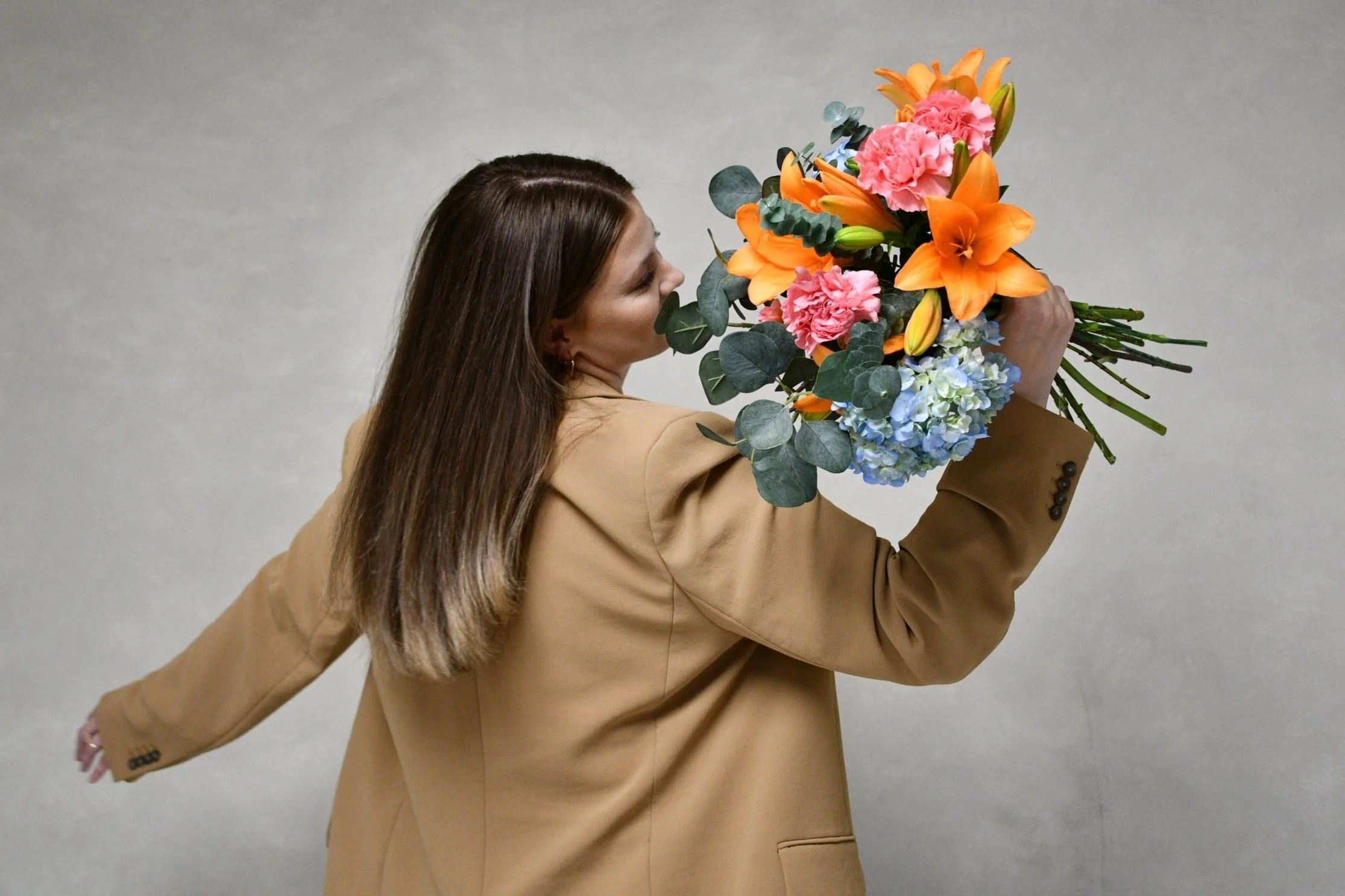 A woman with long brown hair wearing a beige blazer holding a large bouquet of colorful flowers.