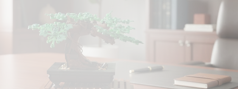 A miniature LEGO bonsai tree with green leaves and a brown trunk on a wooden desk with a pen and a closed notebook nearby.