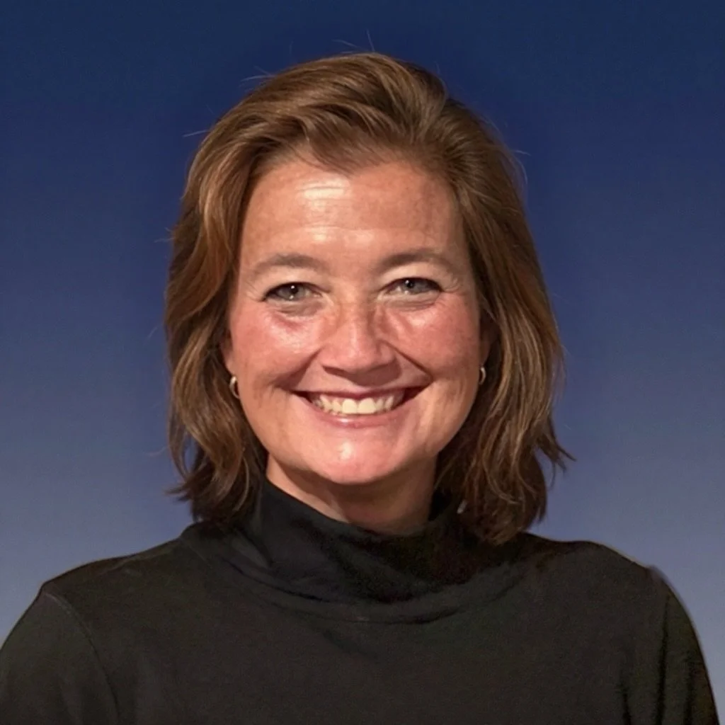A woman with shoulder-length reddish-brown hair smiling, wearing a black turtleneck against a dark blue background.