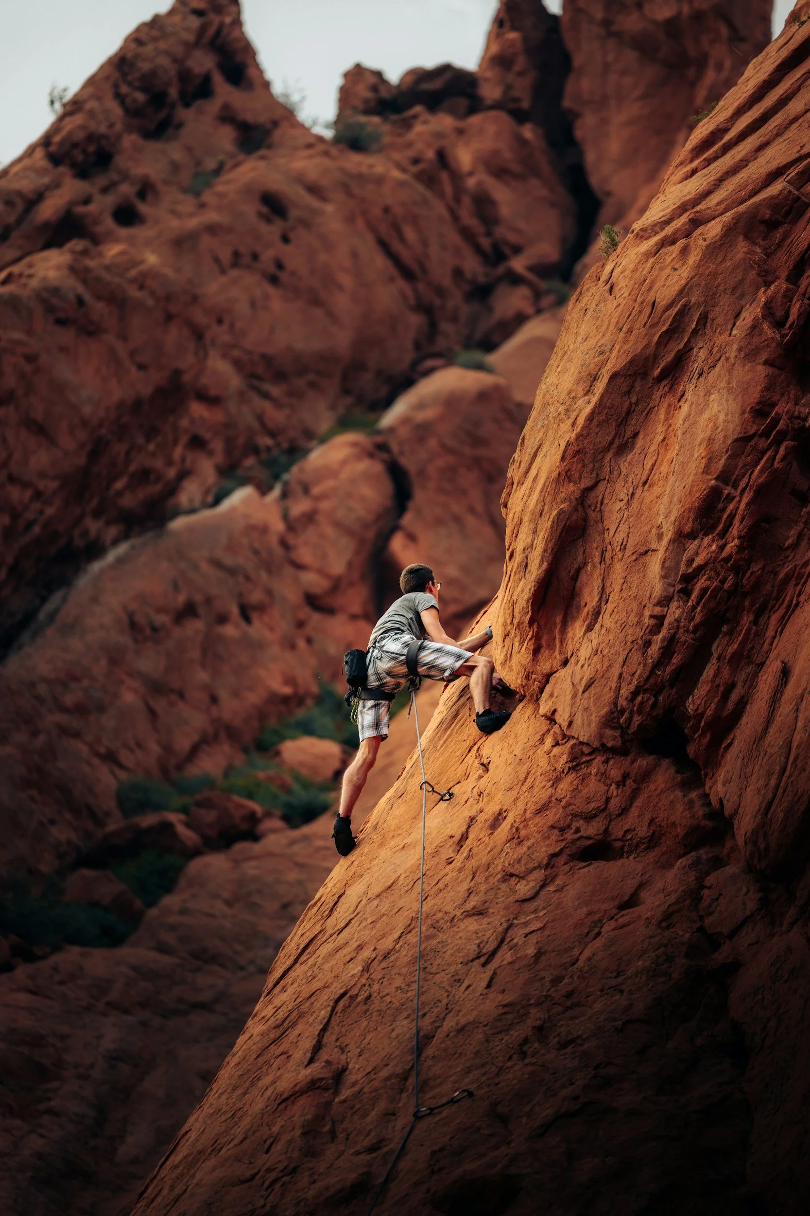 A person rock climbing on a red sandstone cliff, wearing a harness and shorts, while holding onto the rock face with one hand.