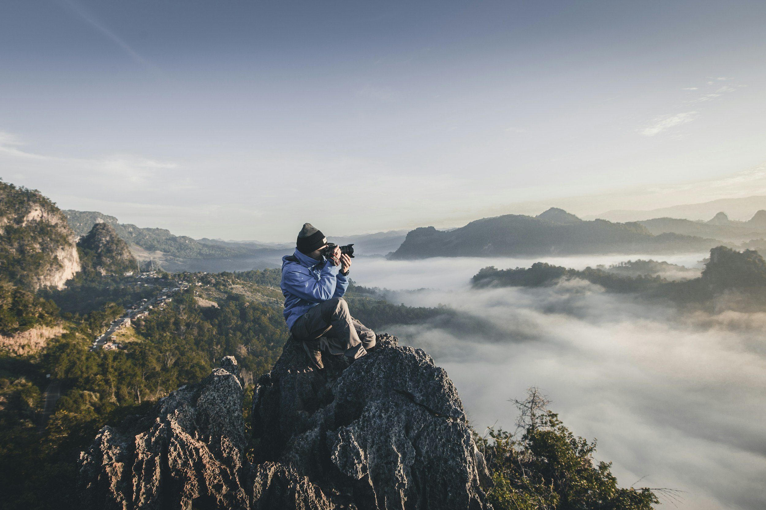 A person in outdoor gear sitting on a rock, taking a photograph of a mountain landscape with fog over the valleys.
