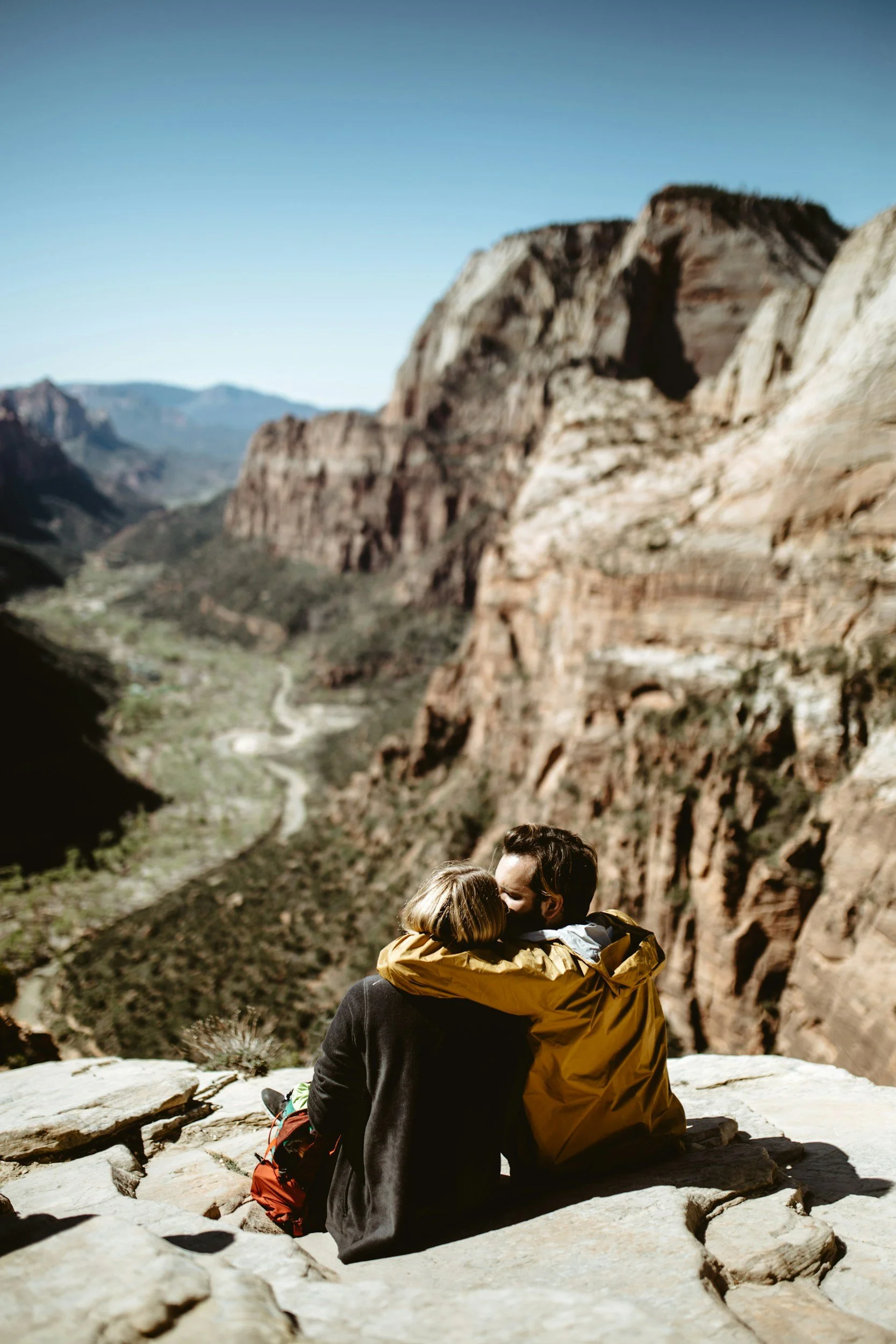 A couple sits embracing on a rocky ledge overlooking a canyon with steep cliffs and a winding river.