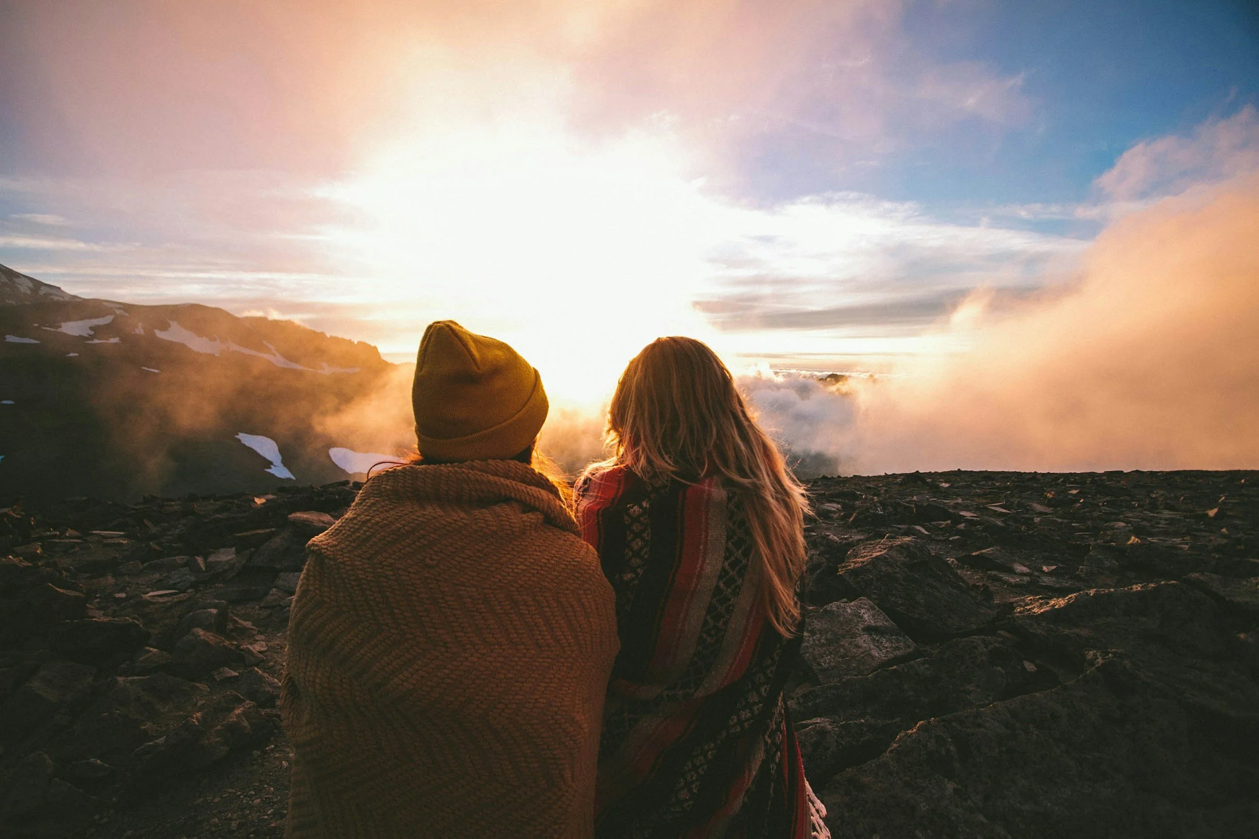 Two people sitting on rocky ground watching a sunset over mountains with clouds and snow patches.
