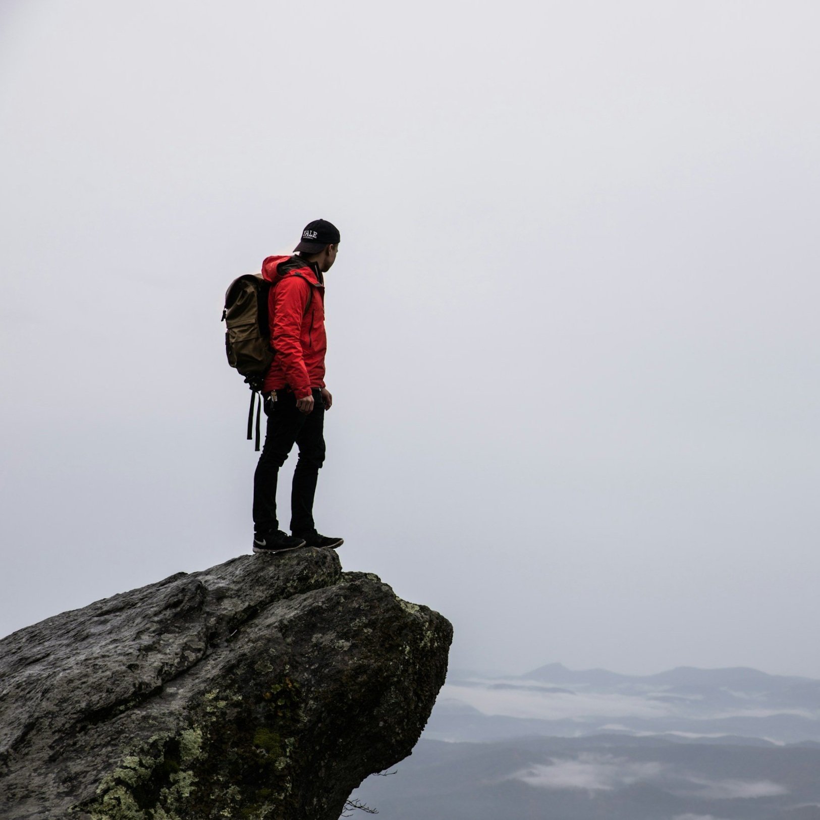A person in a red jacket with a backpack standing on the edge of a large rock formation, looking out over foggy mountains.