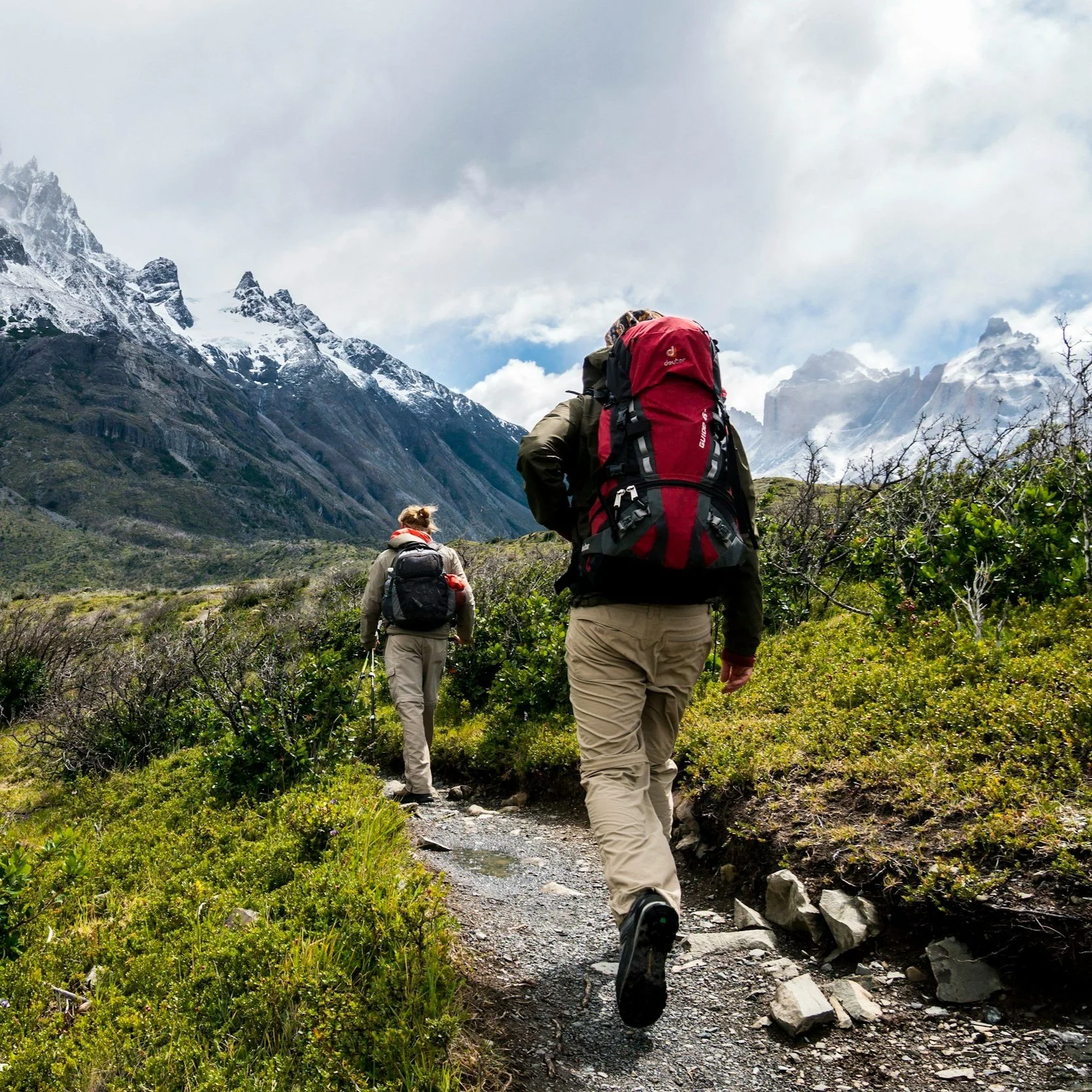 Two hikers carrying backpacks walk along a trail through mountainous terrain with snow-capped peaks in the background.