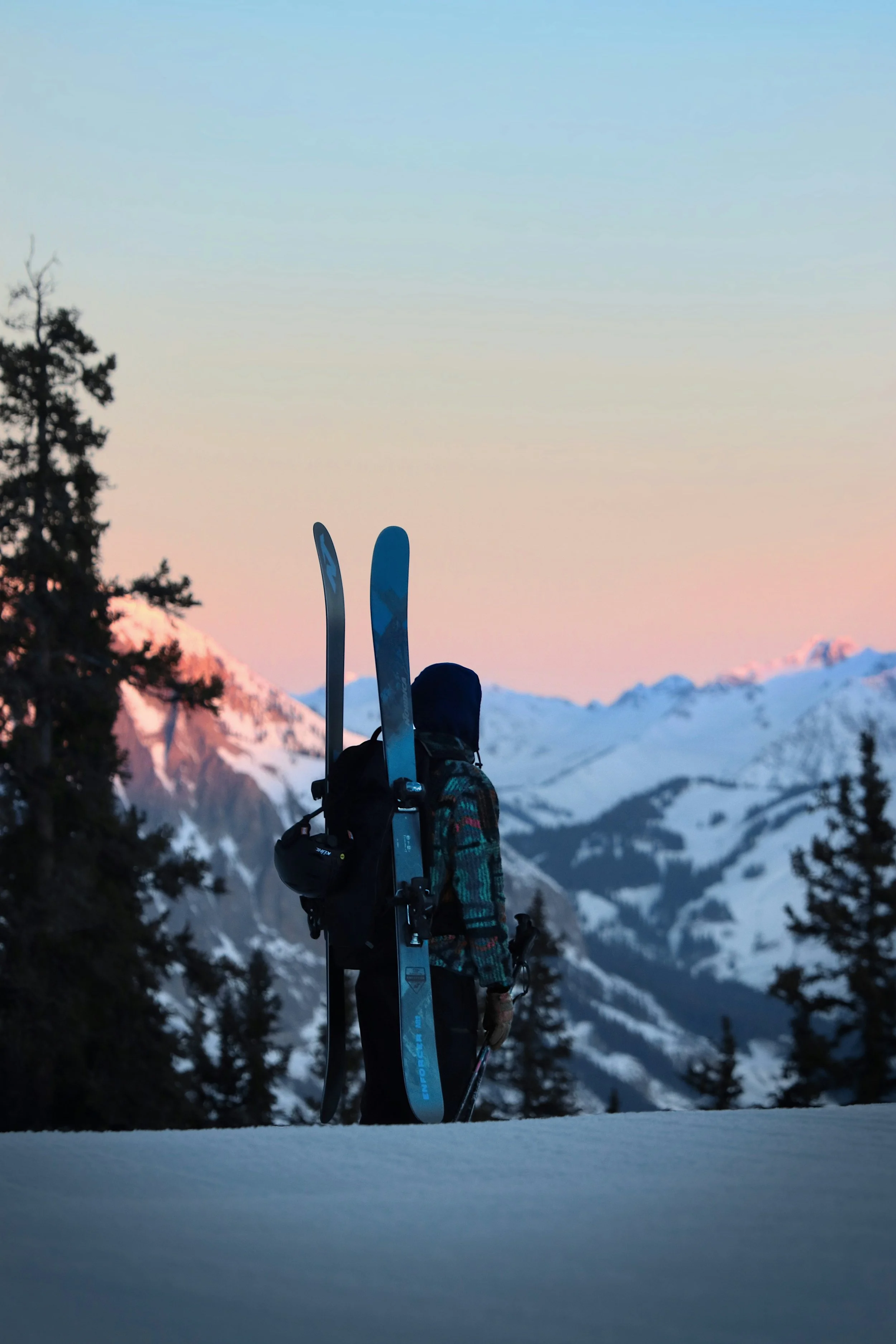 Girl carrying skis in snowy mountains at sunset.