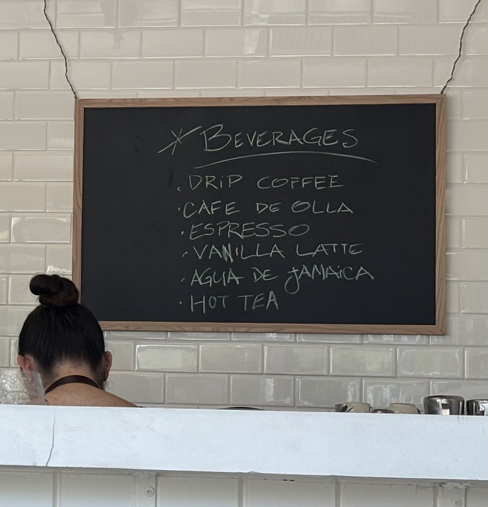 A chalkboard menu listing beverage options including drip coffee, café de olla, espresso, vanilla latte, agua de jamaica, and hot tea, with a person partially visible in the foreground.