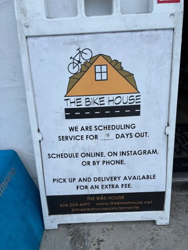 Signboard of The Bike House with a logo of a house and a bicycle on the roof. The sign mentions scheduling service, pick-up and delivery options, and contact information.