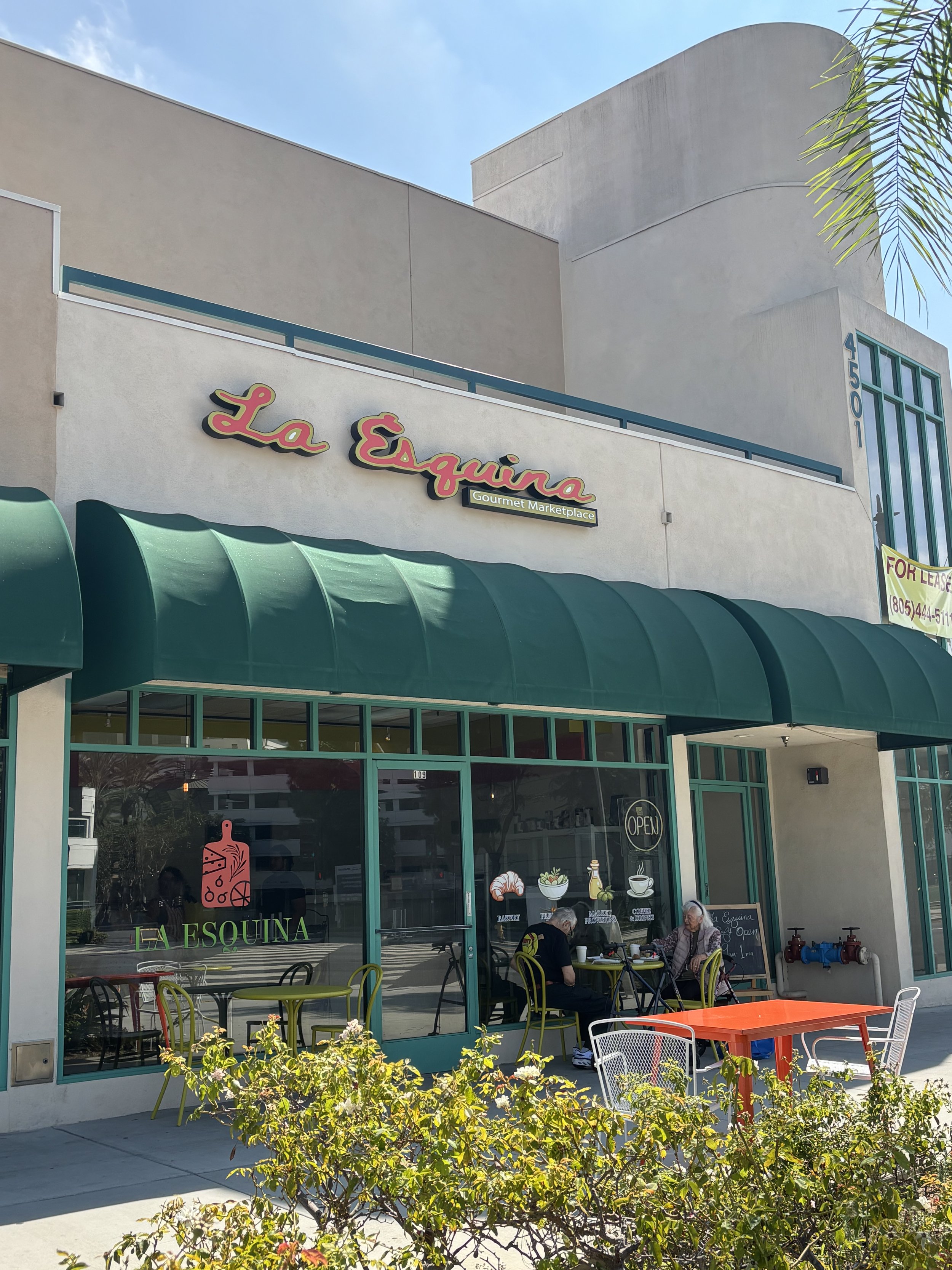 The storefront of La Esquina, a café with outdoor seating in front, surrounded by plants and trees, with a bright blue sky overhead.