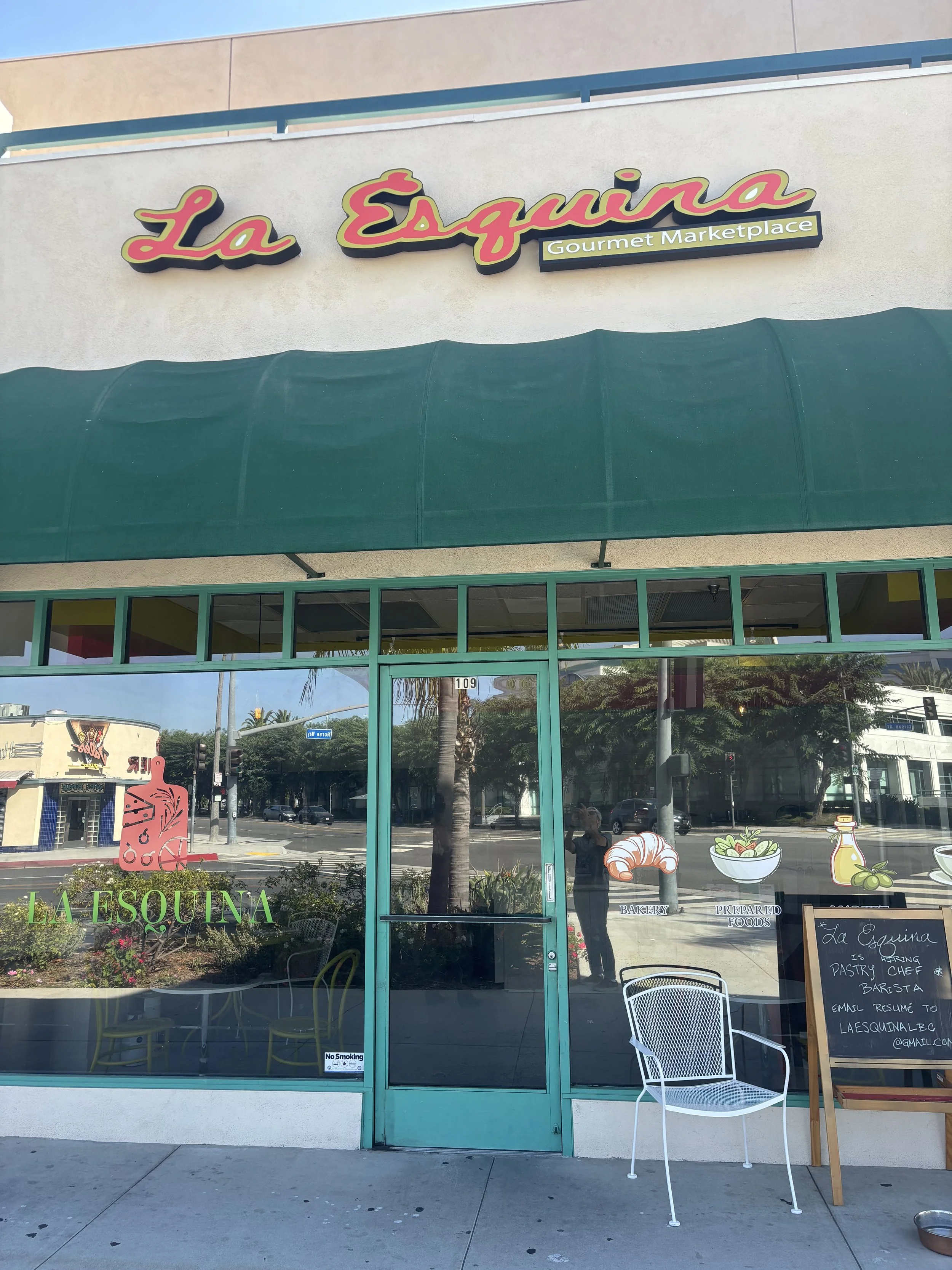 Storefront for La Esquina, a gourmet marketplace and bakery, with a green awning, glass door, and window decals of baked goods and prepared foods, sidewalk seating, and a chalkboard menu.