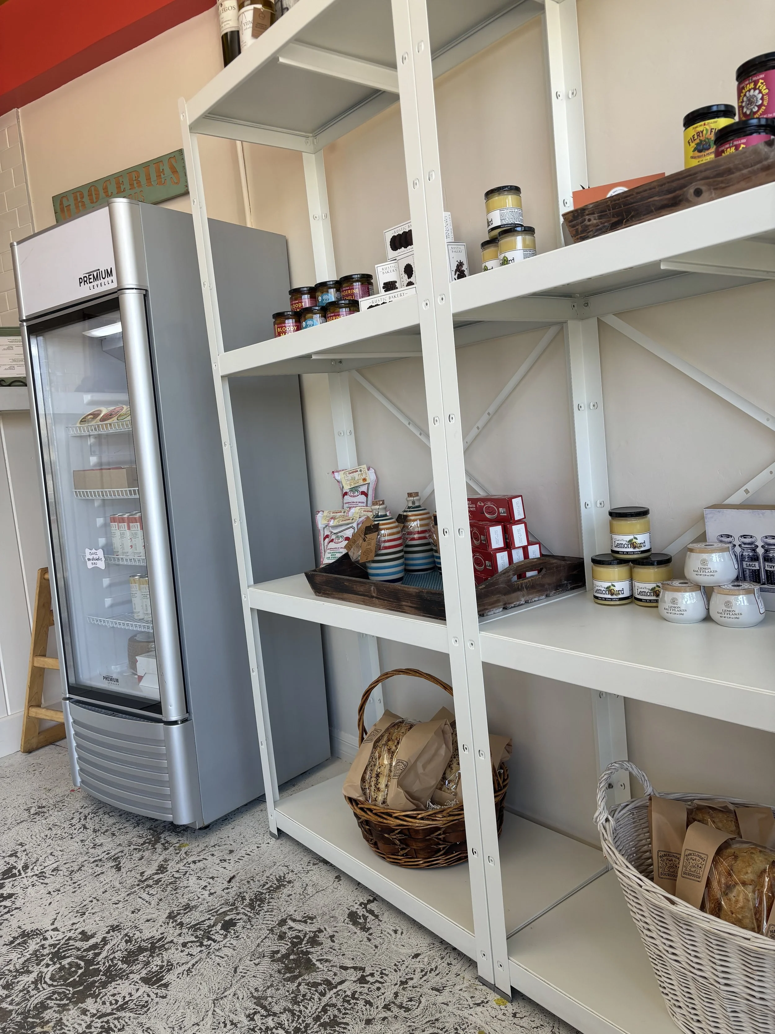 Empty white metal shelving unit with baskets of bread and jars of spreads, standing next to a silver refrigerator in a grocery or café interior.