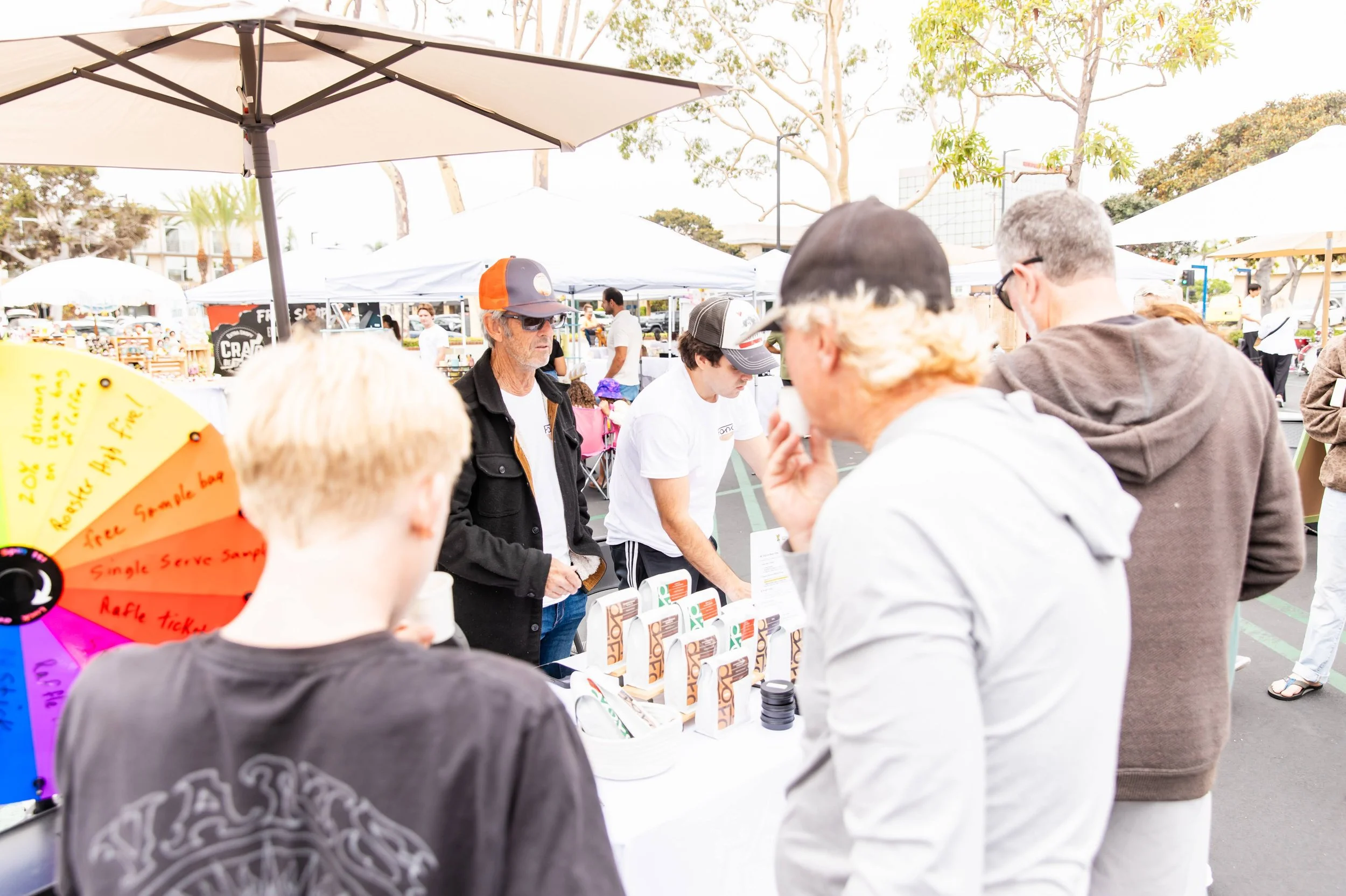 People gathered at an outdoor market booth under large umbrellas, engaging in conversation and browsing products on display, with trees and additional market tents in the background.