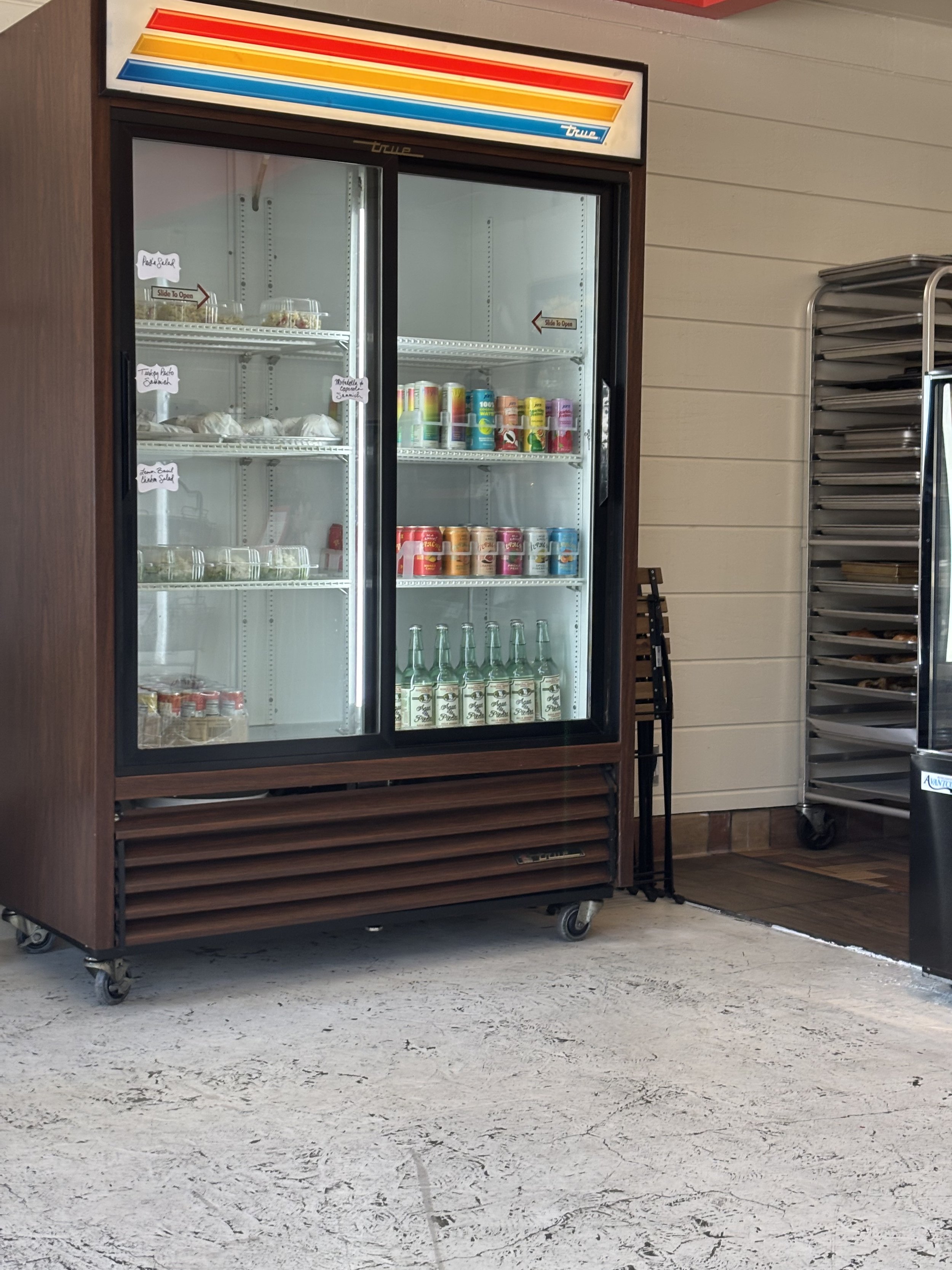 Empty refrigerated display case with glass doors, containing some canned drinks and bottled beverages, situated on a cart with wheels.
