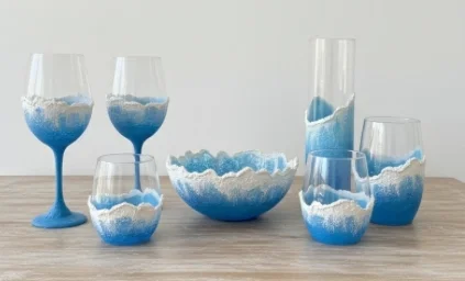 Glassware with blue and white layered sand, including wine glasses, a bowl, and tumblers on a wooden surface.