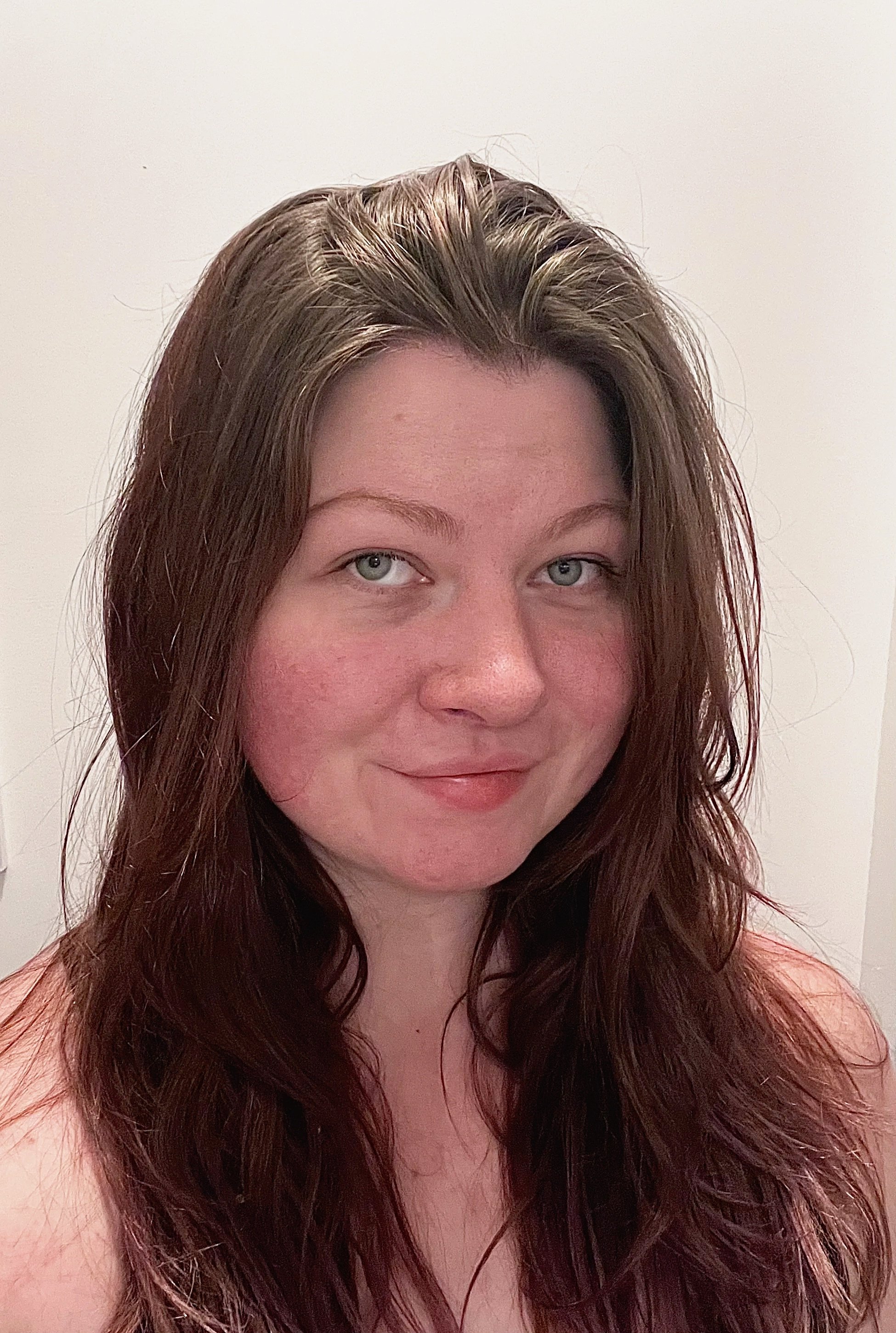 A woman with long, wavy, reddish-brown hair and blue eyes, smiling softly, standing against a plain, light-colored background.