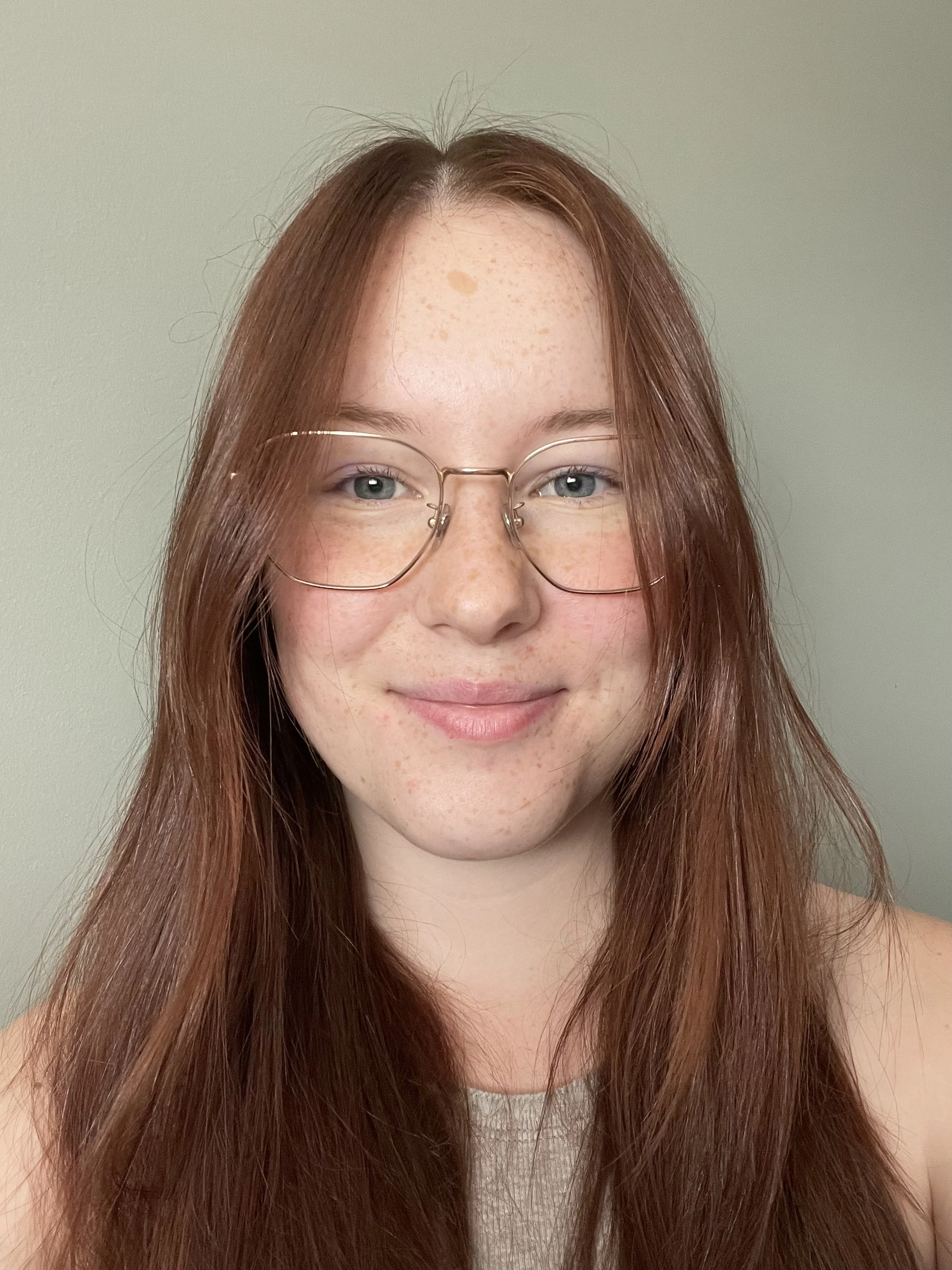 A young woman with long red hair, wearing glasses, smiling softly against a plain light green background.