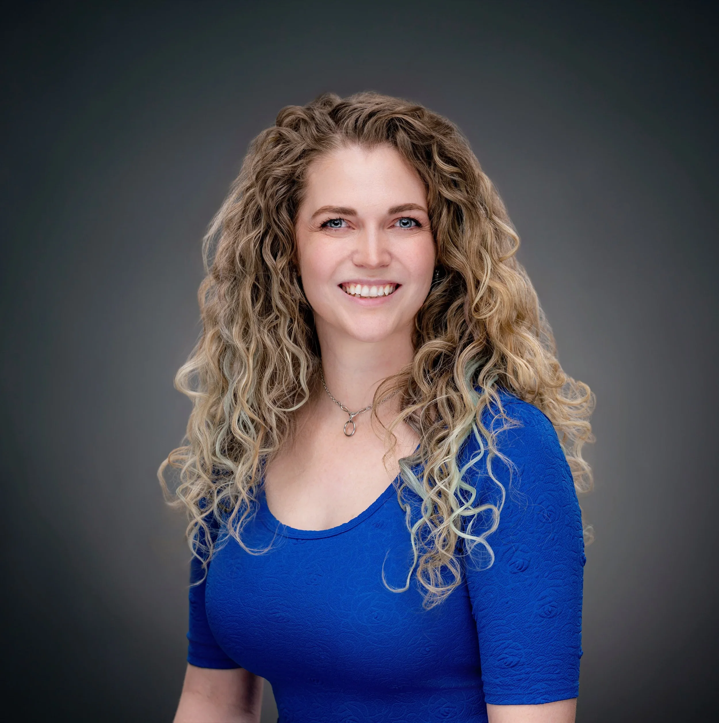 A picture of Ashley,  woman with curly blonde hair and blue eyes smiling, wearing a blue textured dress, against a dark gray background.
