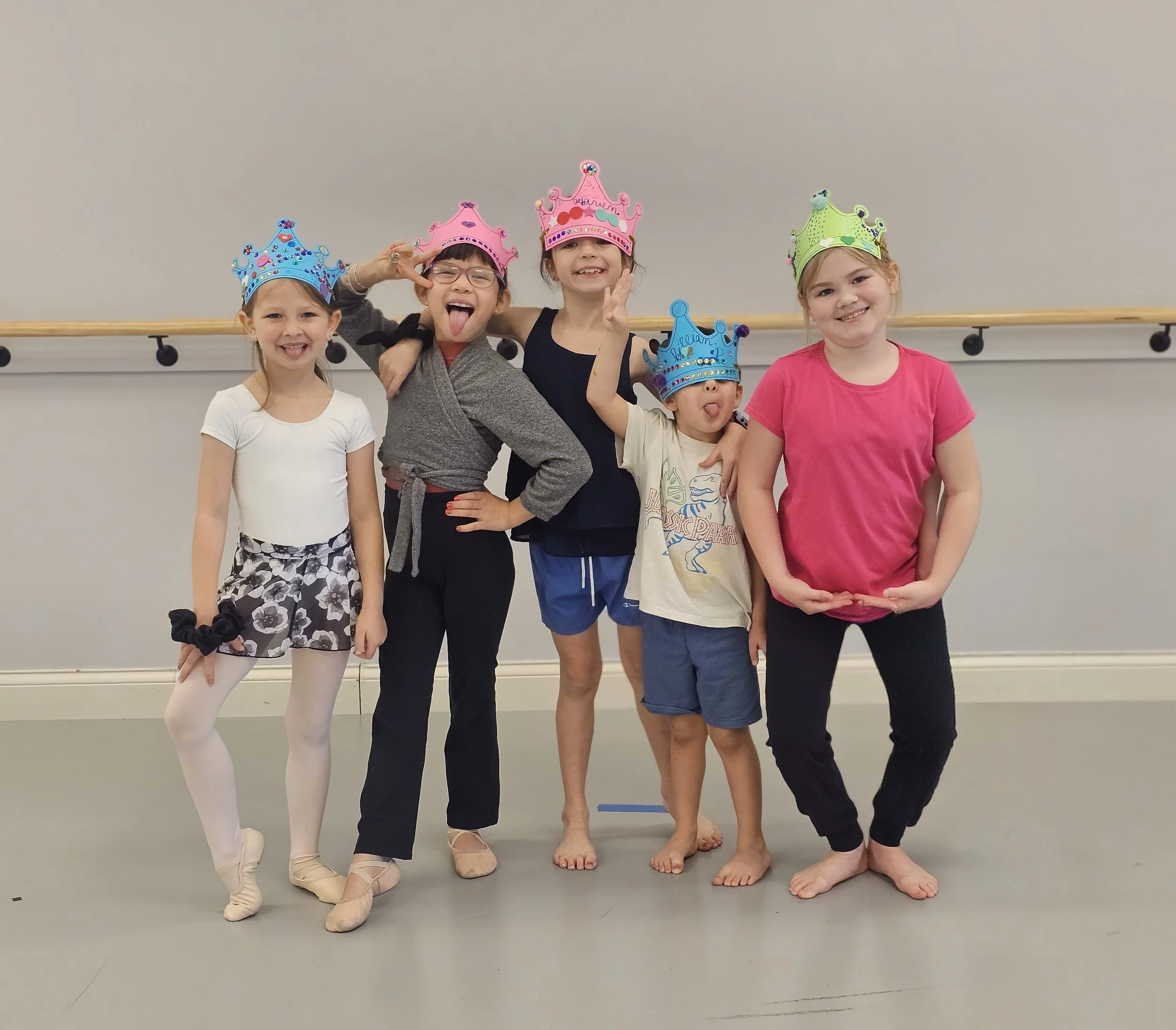 Group of five children in ballet costumes and colorful crowns standing in a dance studio with a gray wall and ballet barre behind them. They are smiling, making funny faces, and celebrating.