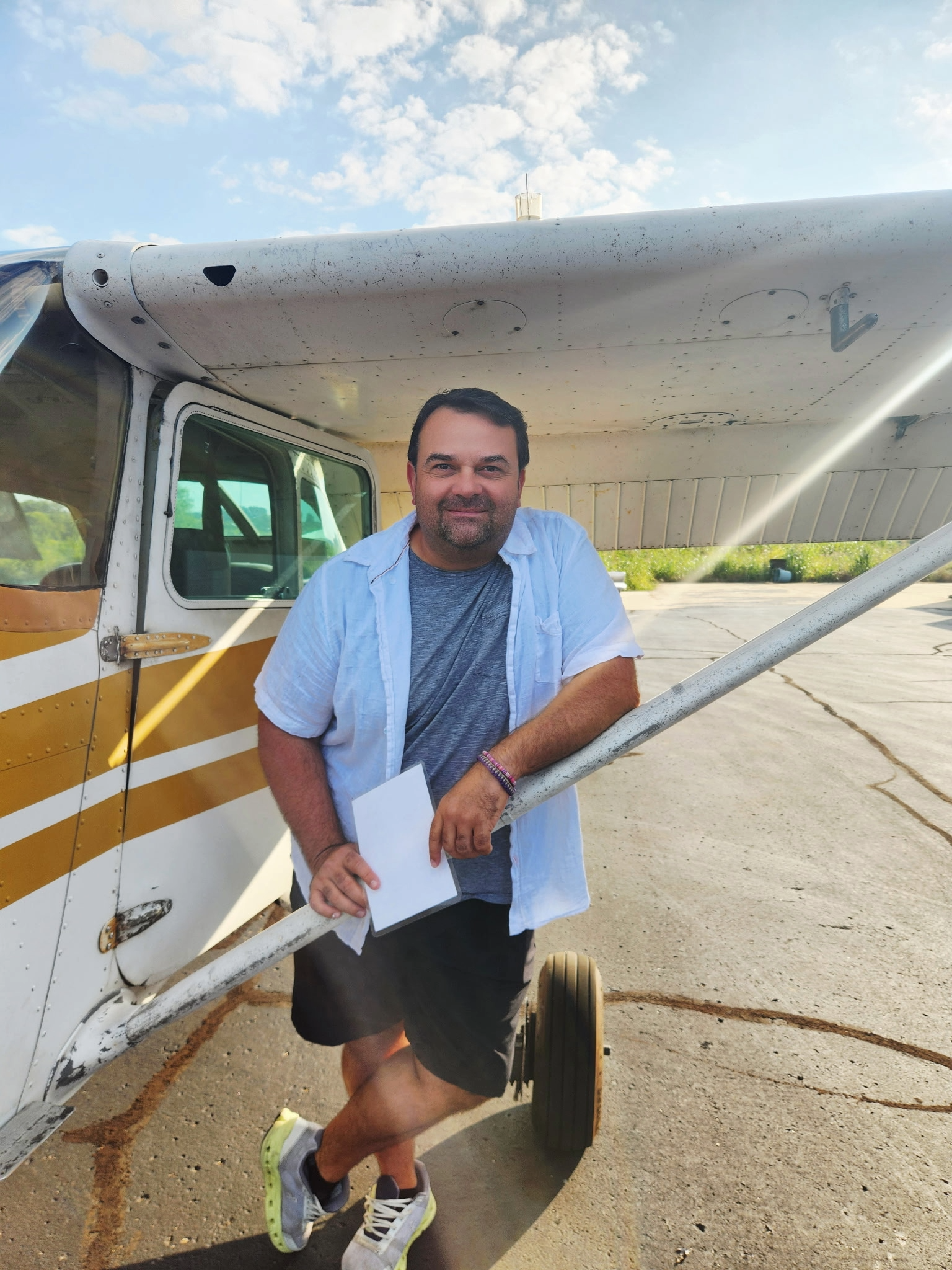A man standing in front of a small airplane with a smile on his face. The airplane is white with gold stripes and is parked on a concrete surface. The sky is partly cloudy with blue patches.